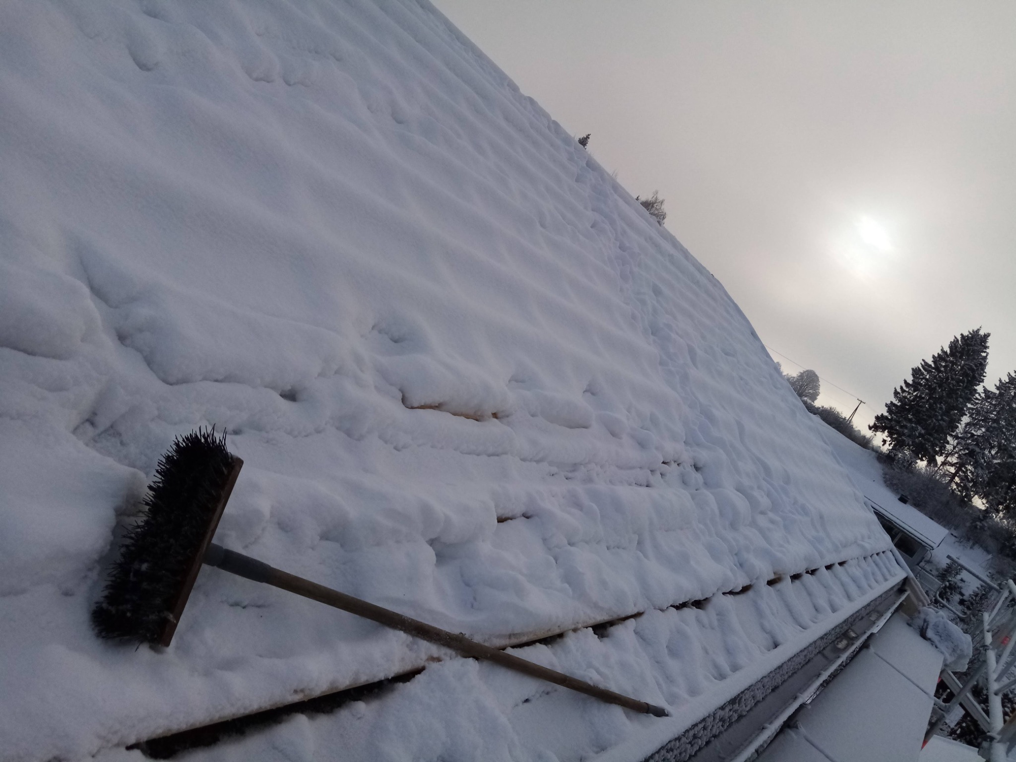 Śnieżny dach z widocznymi śladami stóp i szczotką do odśnieżania, pochmurne niebo w tle.