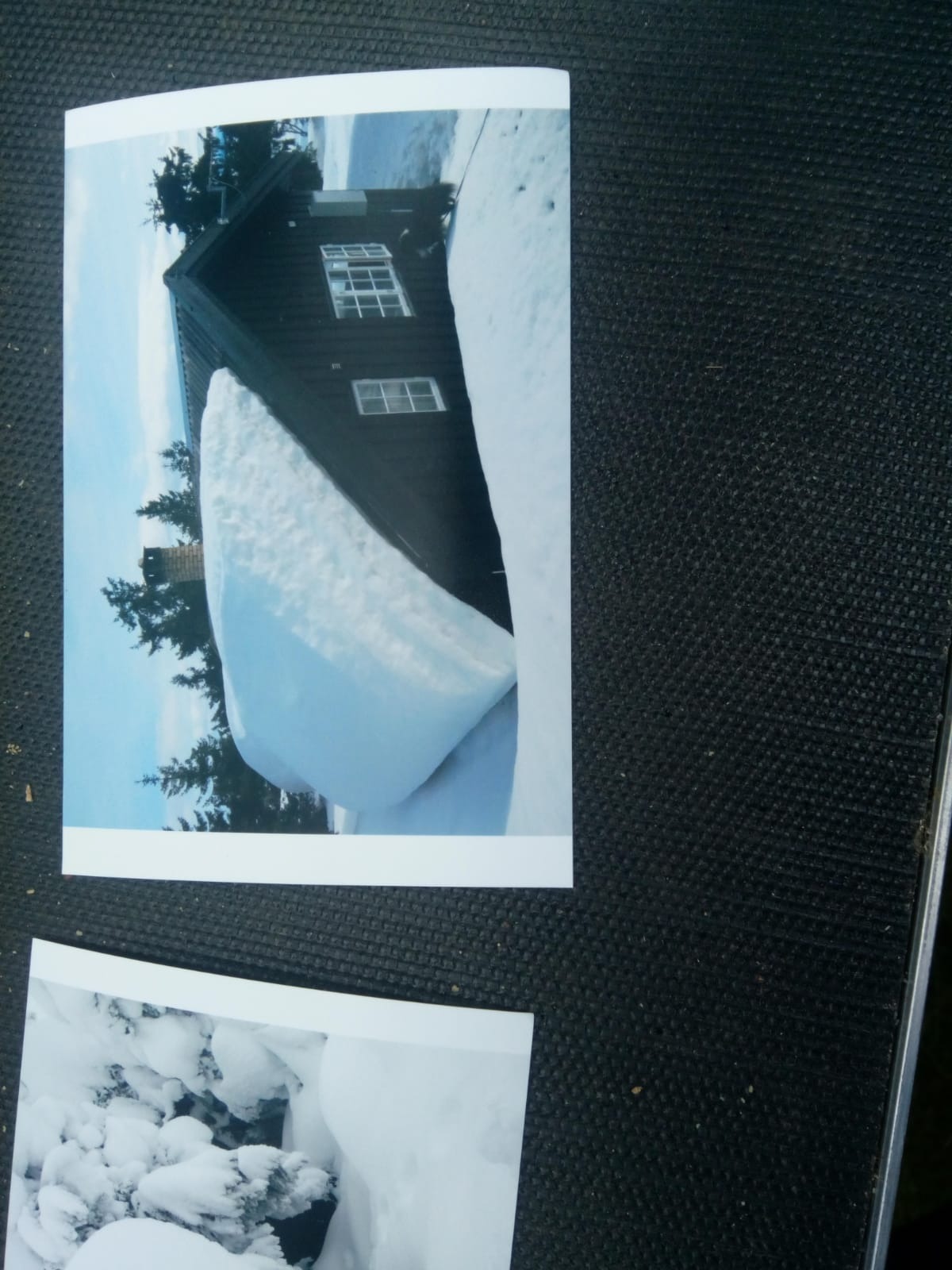 Zdjęcie domu z dachem pokrytym grubą warstwą śniegu, częściowo wiszącego nad krawędzią, w tle drzewa iglaste i błękitne niebo. Na dole kadru widoczny fragment innego zdjęcia przedstawiającego śnieg.