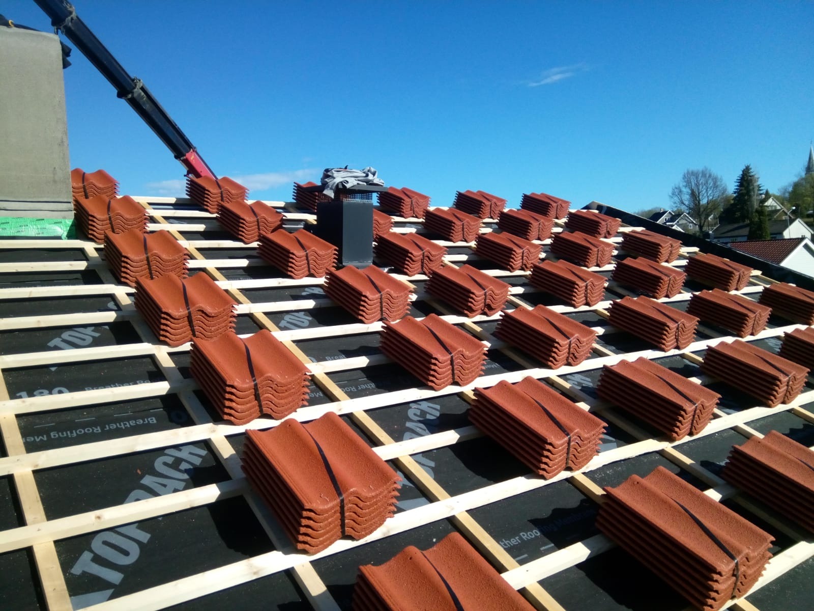 Układanie brązowej dachówki ceramicznej na łatach na dachu pokrytym membraną, widoczny dźwig z podnośnikiem, błękitne niebo w tle.