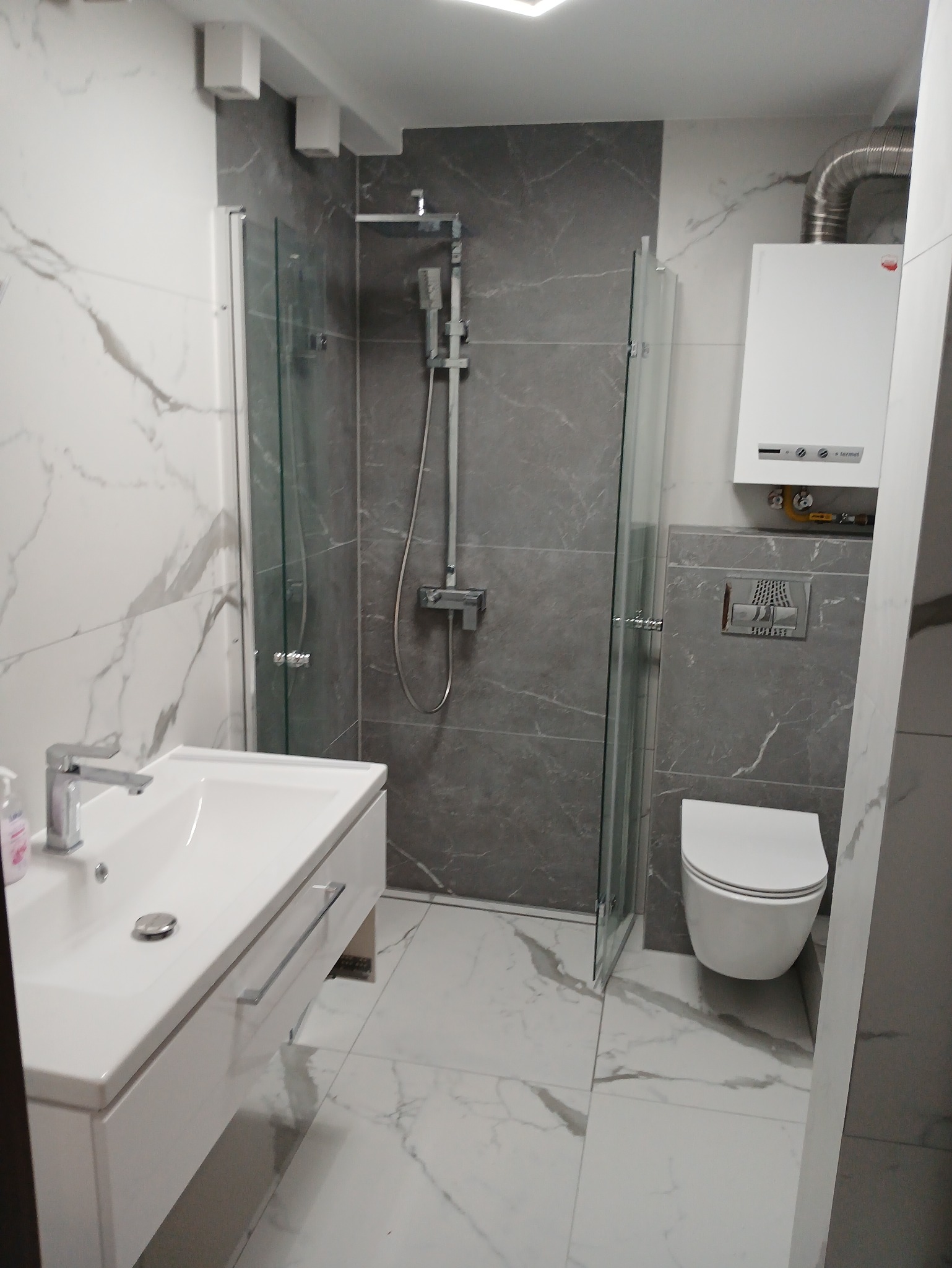 Wykończona łazienka w Kielcach: kabina prysznicowa, umywalka z szafką i toaleta w nowoczesnym stylu. Szare i białe marmurowe płytki na ścianach i podłodze.