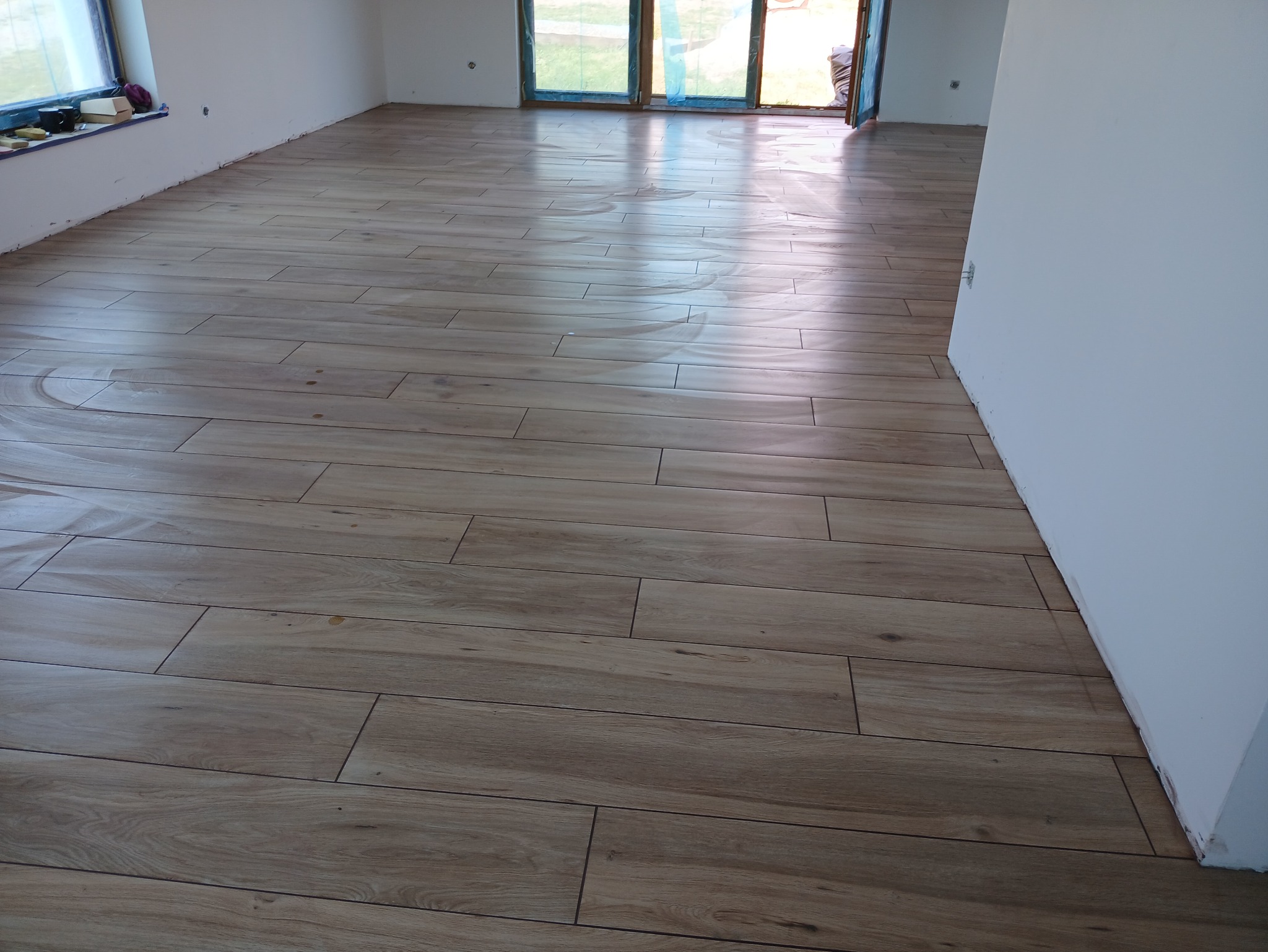 Ułożona podłoga z płytek imitujących drewno w jasnym odcieniu, widoczne świeże fugi i fragmenty białych ścian w nowym wnętrzu. Okno i drzwi balkonowe zabezpieczone folią.