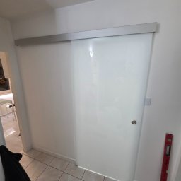 GÓRECKI BUDOWNICTWO Dominik Górecki - Nowoczesne, przesuwne drzwi szklane z aluminiową prowadnicą w jasnym wnętrzu. Po prawej stronie widoczna poziomica, sugerująca precyzyjny montaż.