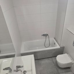 GÓRECKI BUDOWNICTWO Dominik Górecki - Nowoczesna łazienka w odcieniach bieli i szarości. Umywalka z szafką, wanna obudowana szarą płytką, podwieszana toaleta i duże, jasne płytki na ścianach.