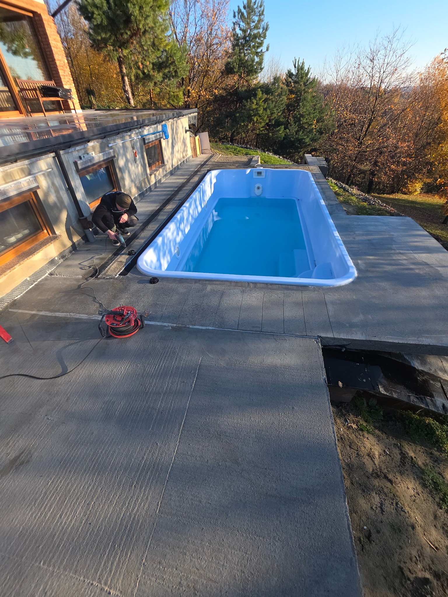 Montaż basenu na betonowym tarasie. Widoczny pracownik podczas instalacji, narzędzia i błękitna woda w basenie. Tło stanowią drzewa i błękitne niebo.