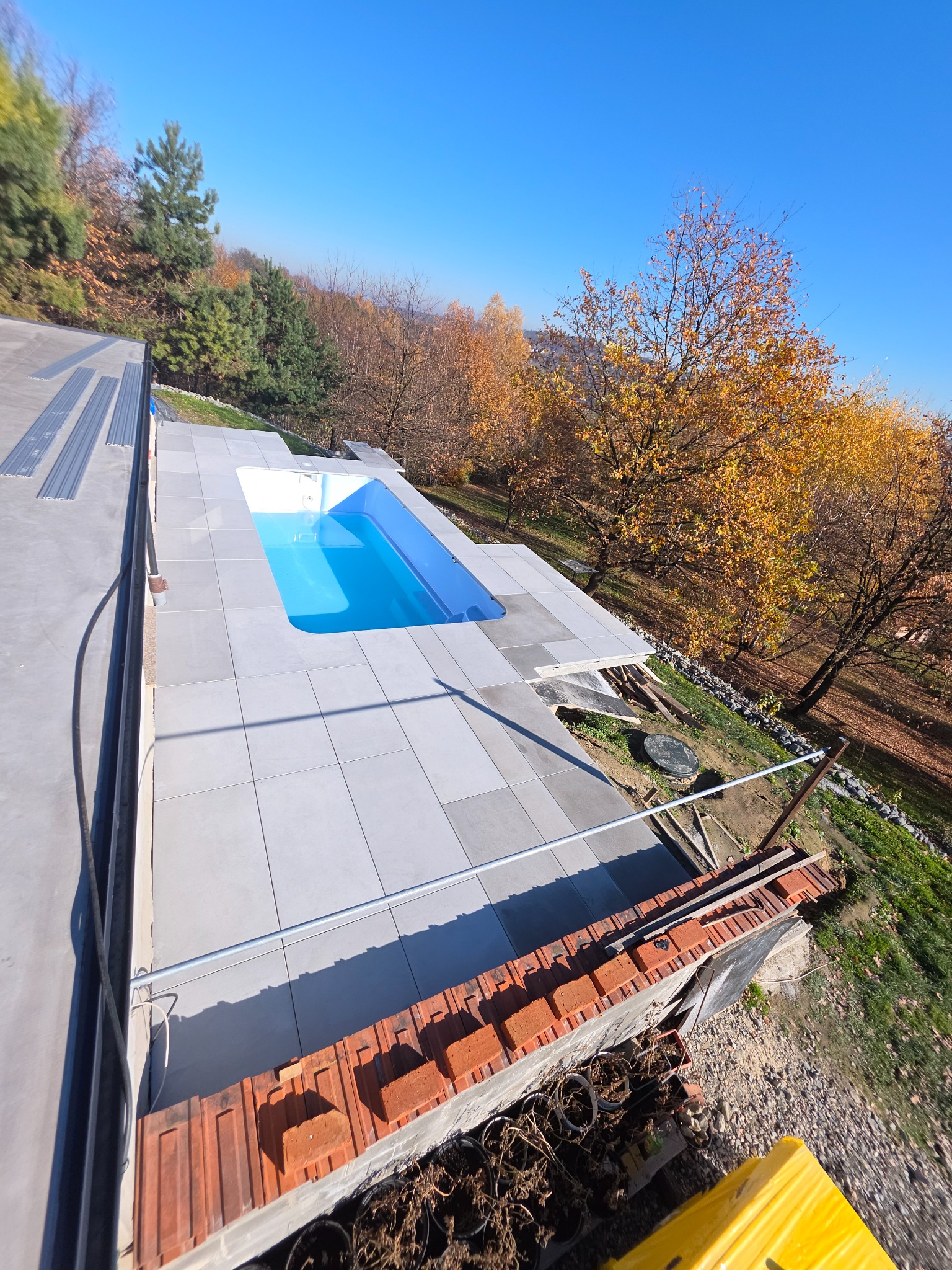 Błękitny basen na tarasie wyłożonym szarą płytką, otoczony jesiennym krajobrazem. Ujęcie z góry, podkreślające geometrię płytek i kontrast kolorów.