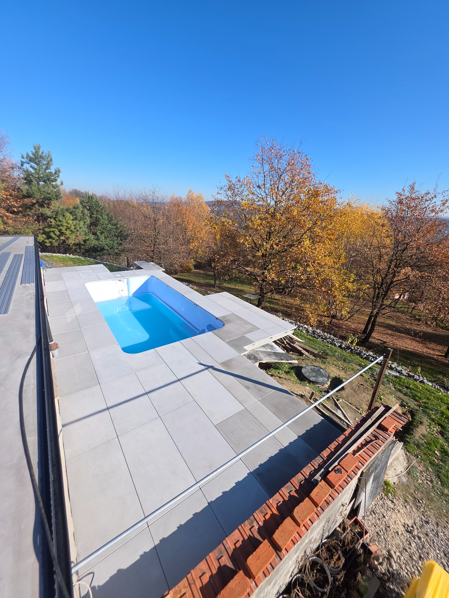 Błękitny basen z szarymi płytkami na tarasie, jesienne drzewa w tle, słoneczny dzień. Widok z góry ukazuje detale wykończenia i otoczenie.