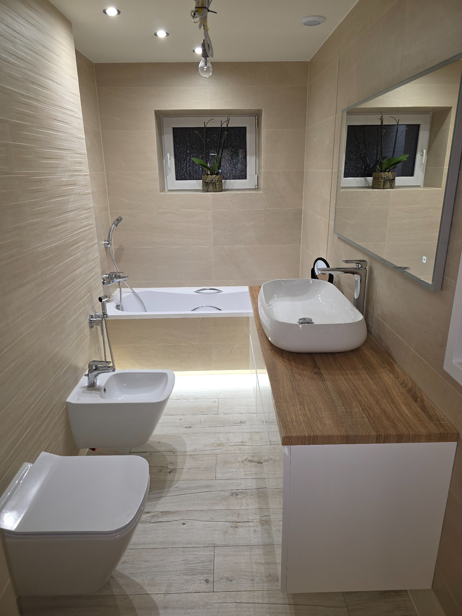 Jasna łazienka z beżowymi płytkami, drewnianym blatem pod umywalką i nowoczesną armaturą. Widoczna wanna, bidet i toaleta w zabudowie.