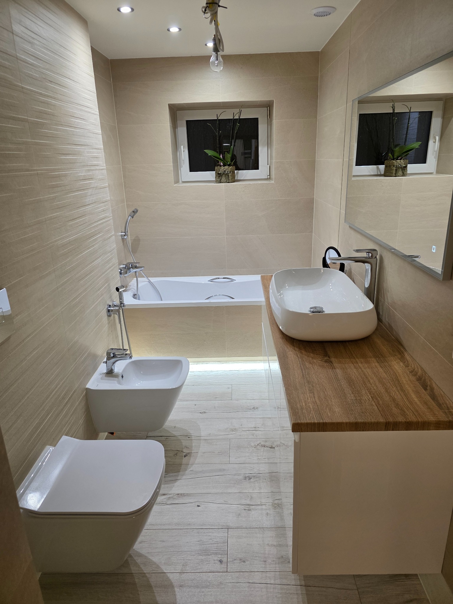 Jasna łazienka po remoncie: beżowe płytki, drewniany blat, biała umywalka, toaleta i bidet. Podświetlana wanna, okno z rośliną. Nowoczesny design.