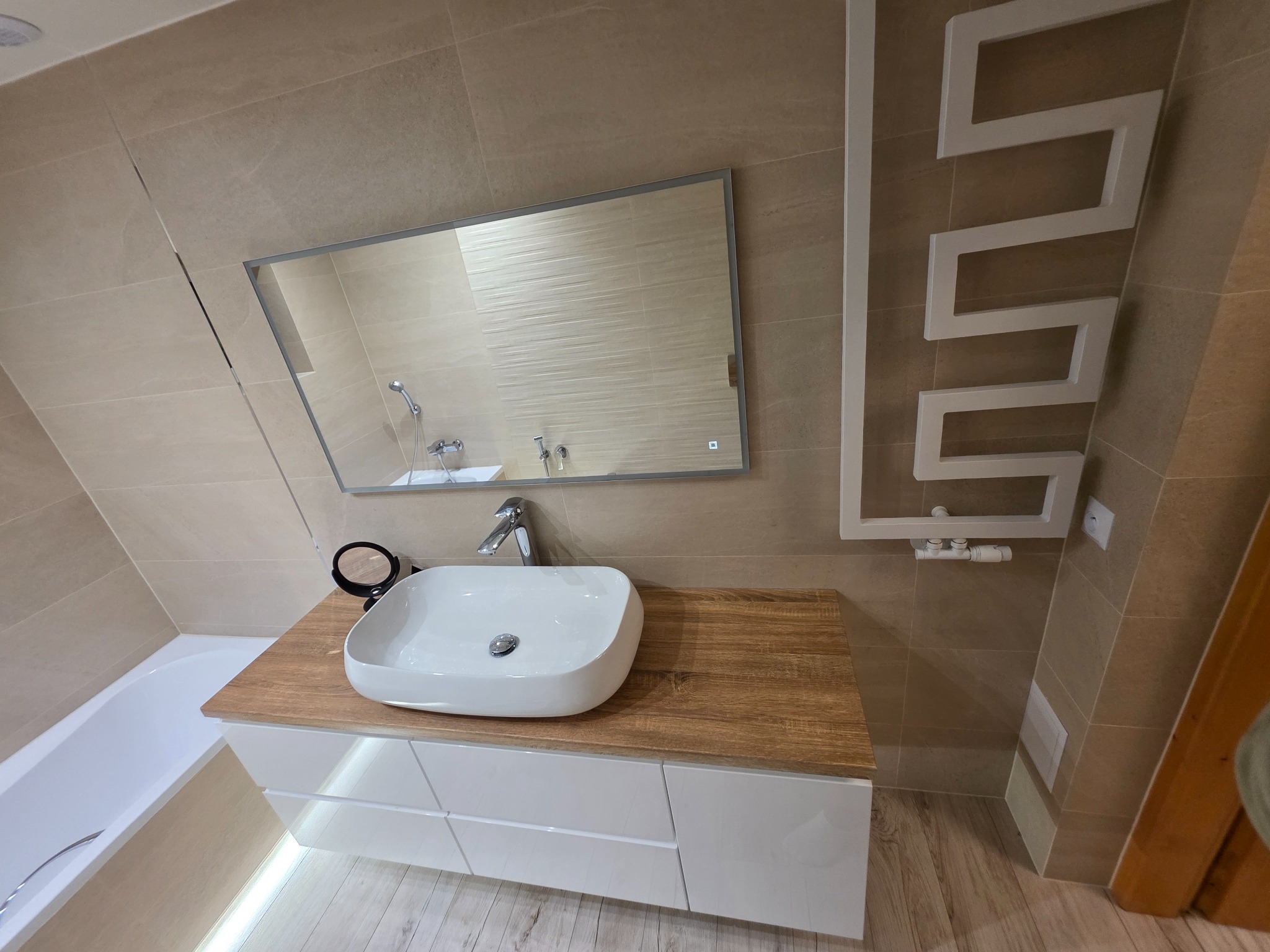 Nowoczesna łazienka z beżowymi płytkami, białą umywalką nablatową na szafce z drewnianym blatem i grzejnikiem drabinkowym. Lustro z podświetleniem LED.