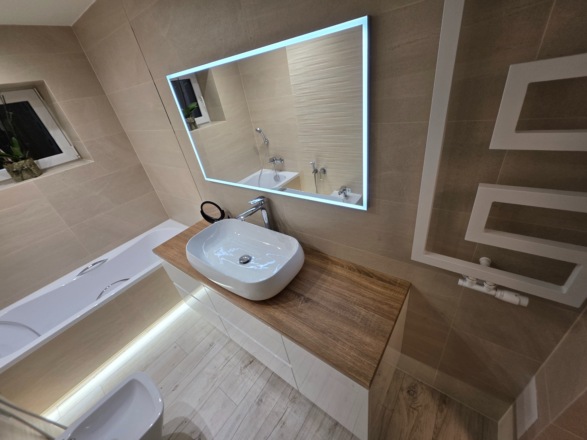 Nowoczesna łazienka z podświetlanym lustrem, białą umywalką na drewnianym blacie i grzejnikiem drabinkowym. Beżowe płytki na ścianach i podłodze.