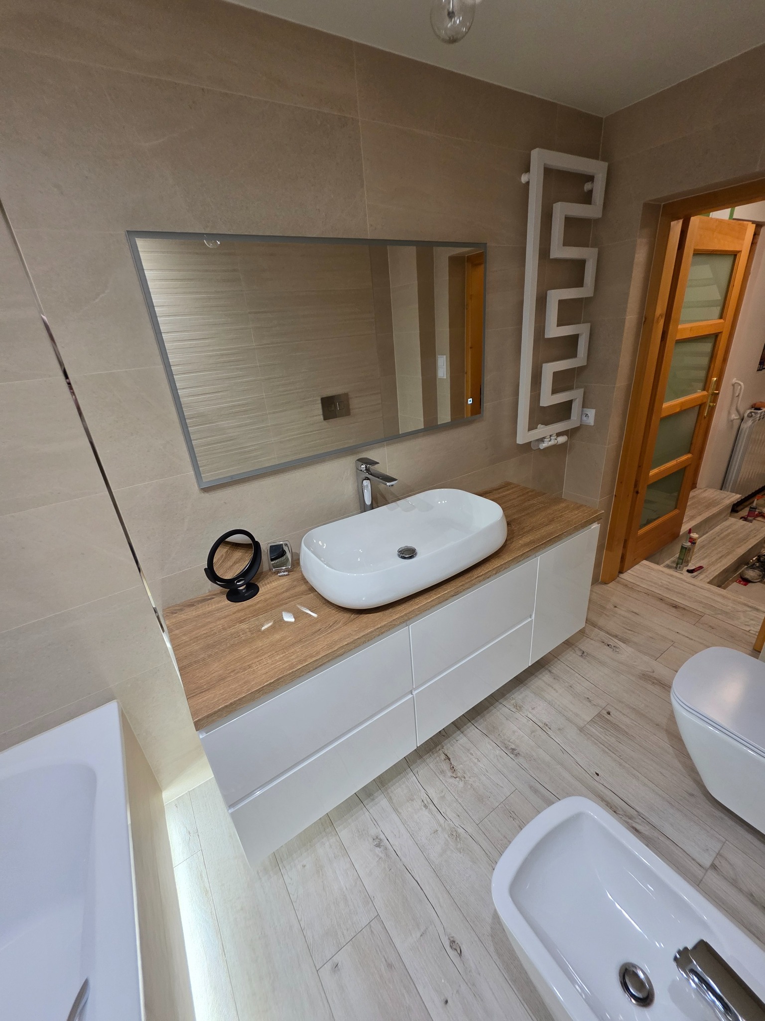 Nowoczesna łazienka po remoncie: beżowe płytki, biała umywalka nablatowa, lustro w szarej ramie, grzejnik drabinkowy i drewnopodobna podłoga. Widok na otwarte drzwi.