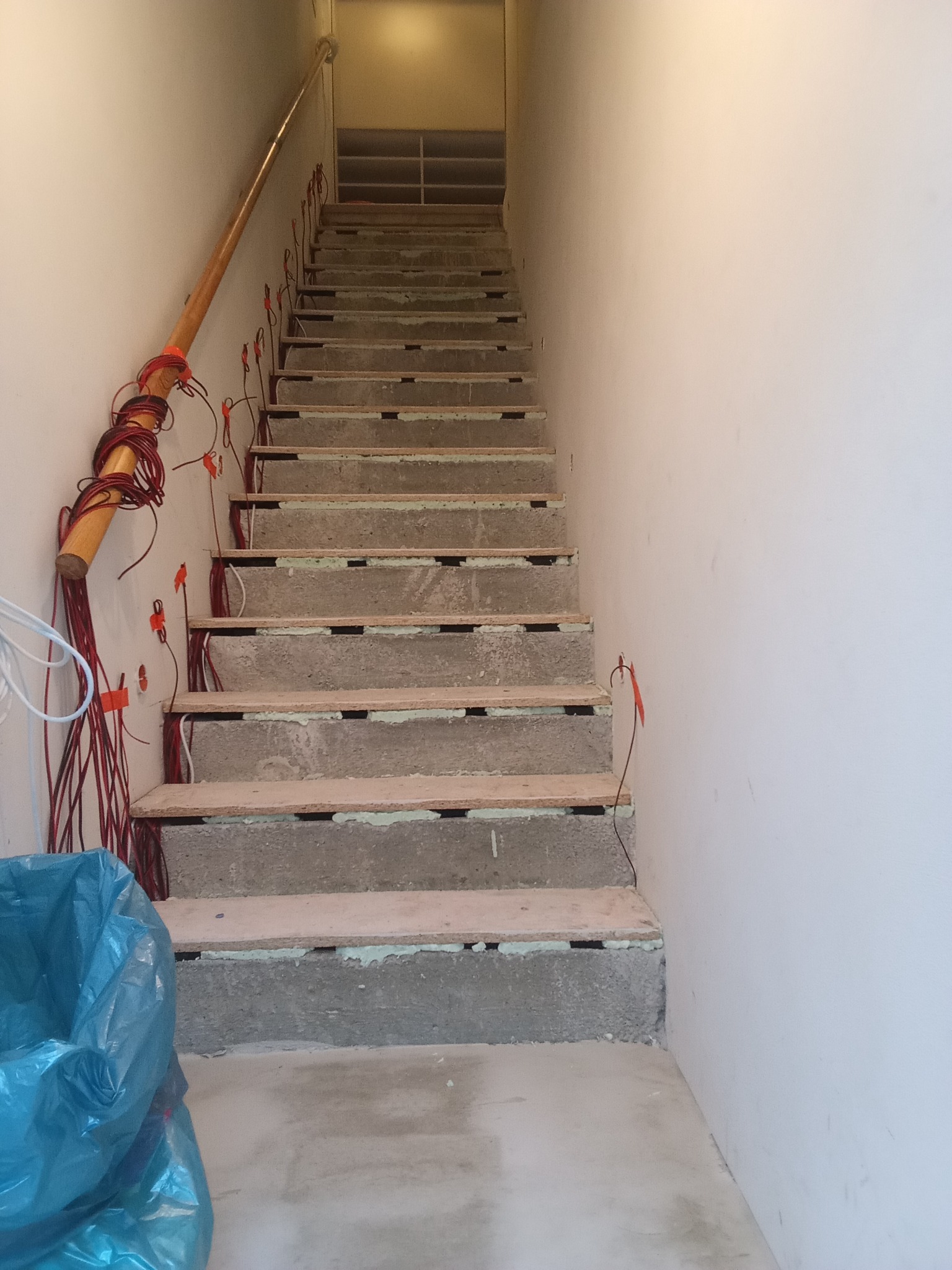Betonowe schody w trakcie montażu, widoczne przewody elektryczne i drewniane elementy. Ujęcie z dołu, perspektywa w górę. W tle widoczne półki.
