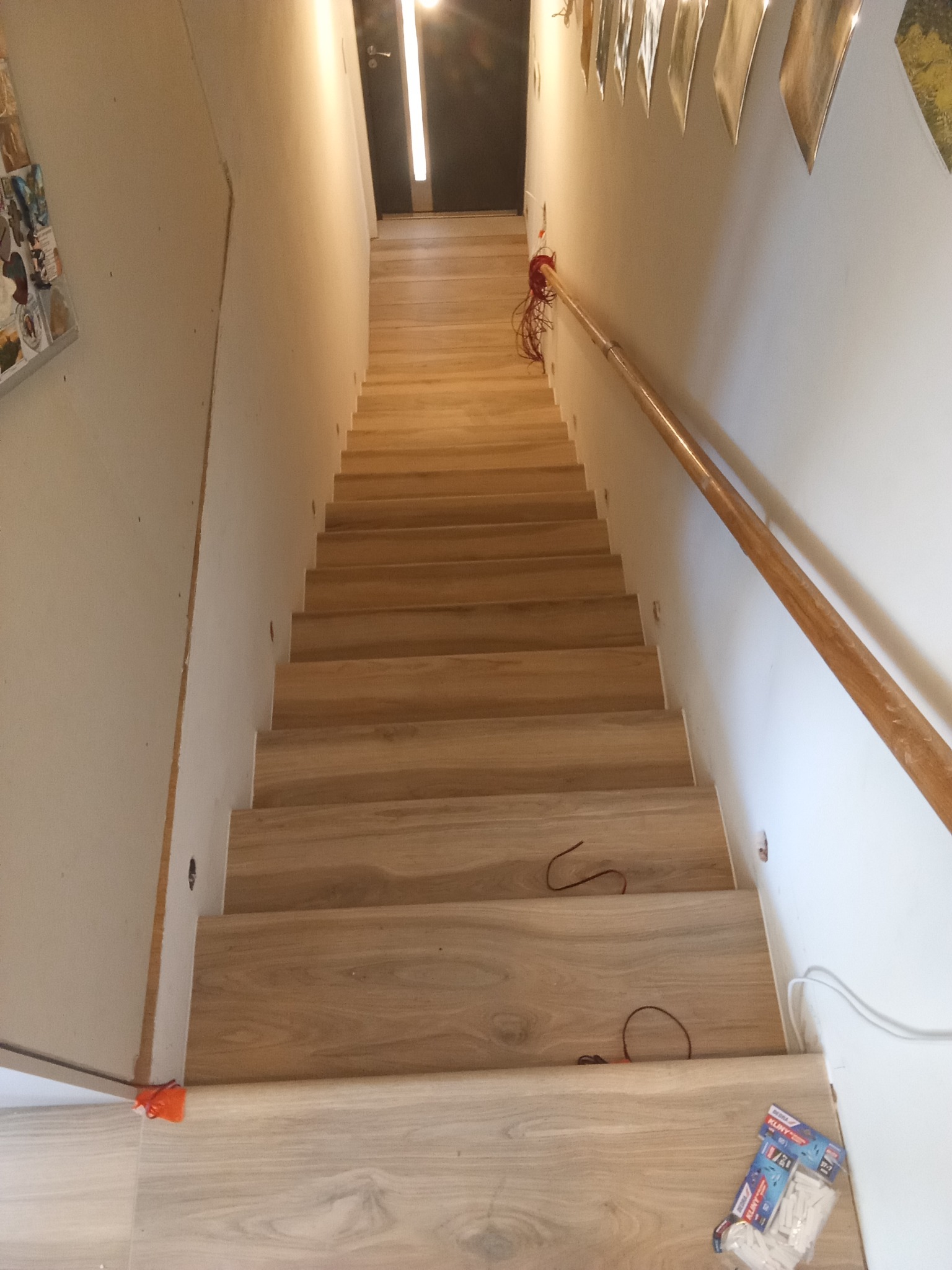 Wykończone schody z drewnopodobnymi panelami, oświetleniem punktowym i drewnianą poręczą. Widoczne kable i akcesoria montażowe.