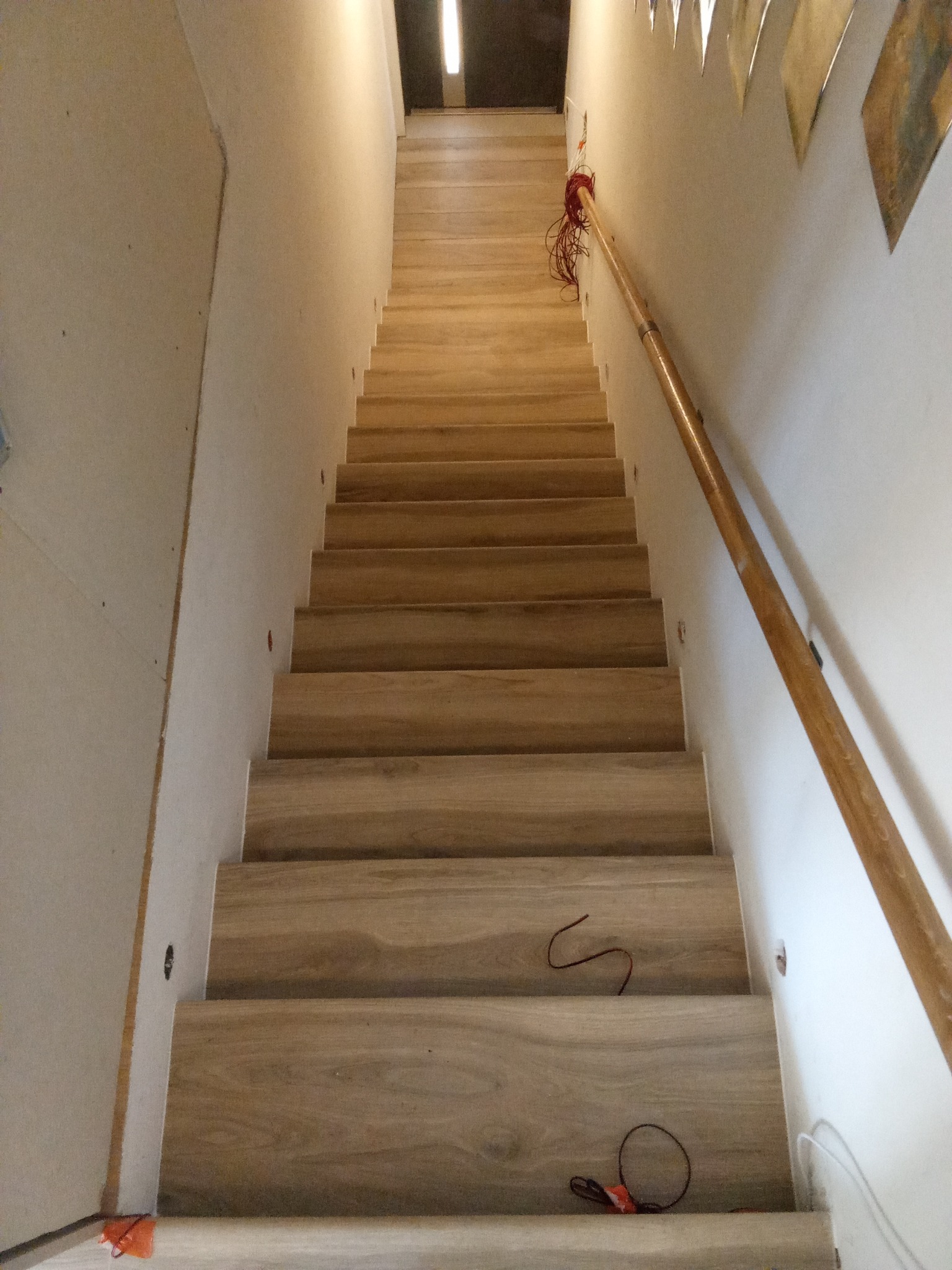 Schody drewniane z oświetleniem punktowym w ścianie, widok z dołu. Poręcz z jasnego drewna po prawej stronie. Widoczne kable elektryczne.