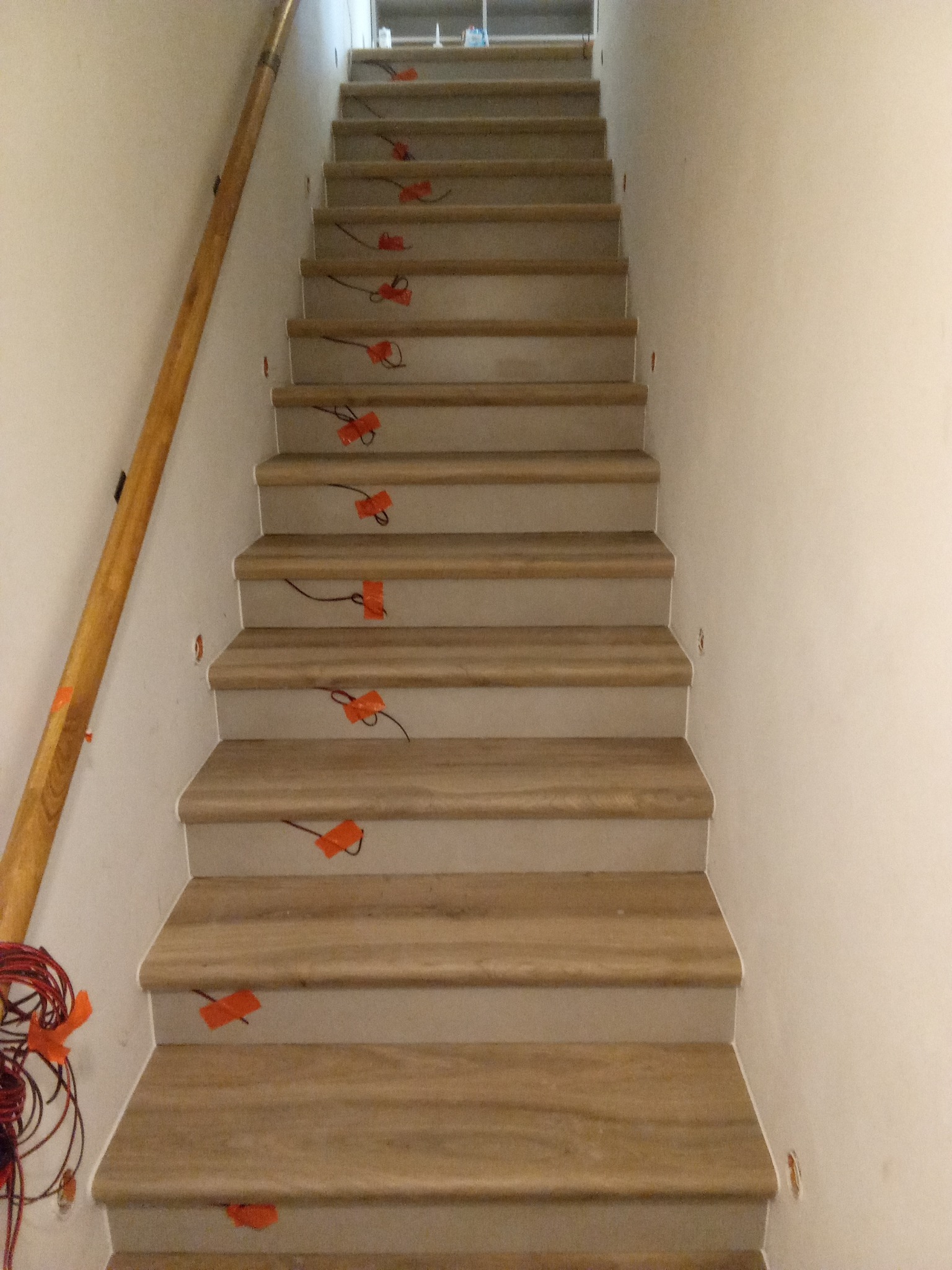 Schody z drewnianymi stopniami w trakcie montażu, zabezpieczone pomarańczową taśmą. Widoczna drewniana poręcz i fragment ściany. Ujęcie z dołu.