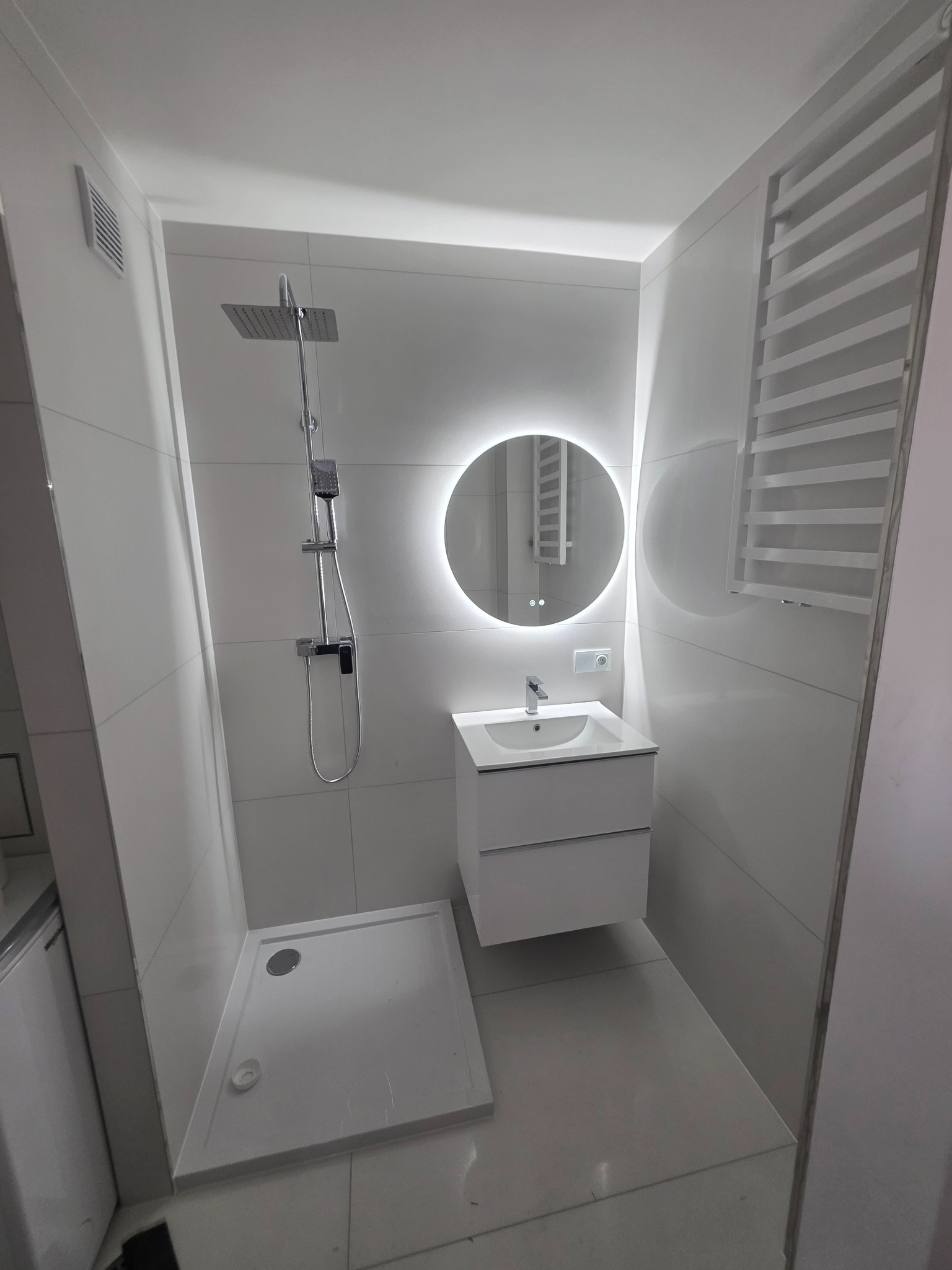 Nowoczesna, minimalistyczna łazienka w bieli: kabina prysznicowa, umywalka z szafką i okrągłe lustro z podświetleniem LED. Grzejnik drabinkowy.