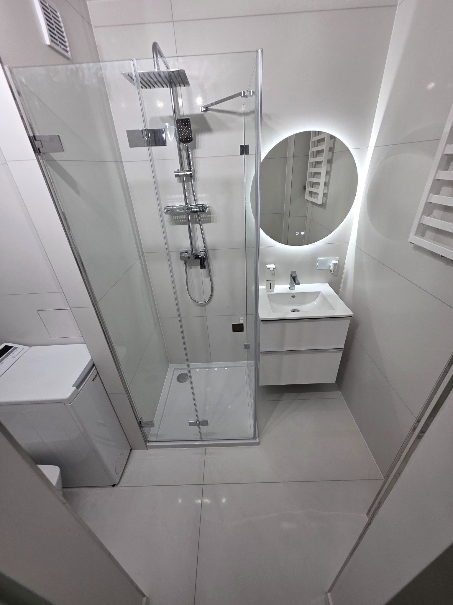 Nowoczesna łazienka w odcieniach szarości z kabiną prysznicową typu walk-in, okrągłym lustrem z podświetleniem LED i pralką. Minimalistyczny design.