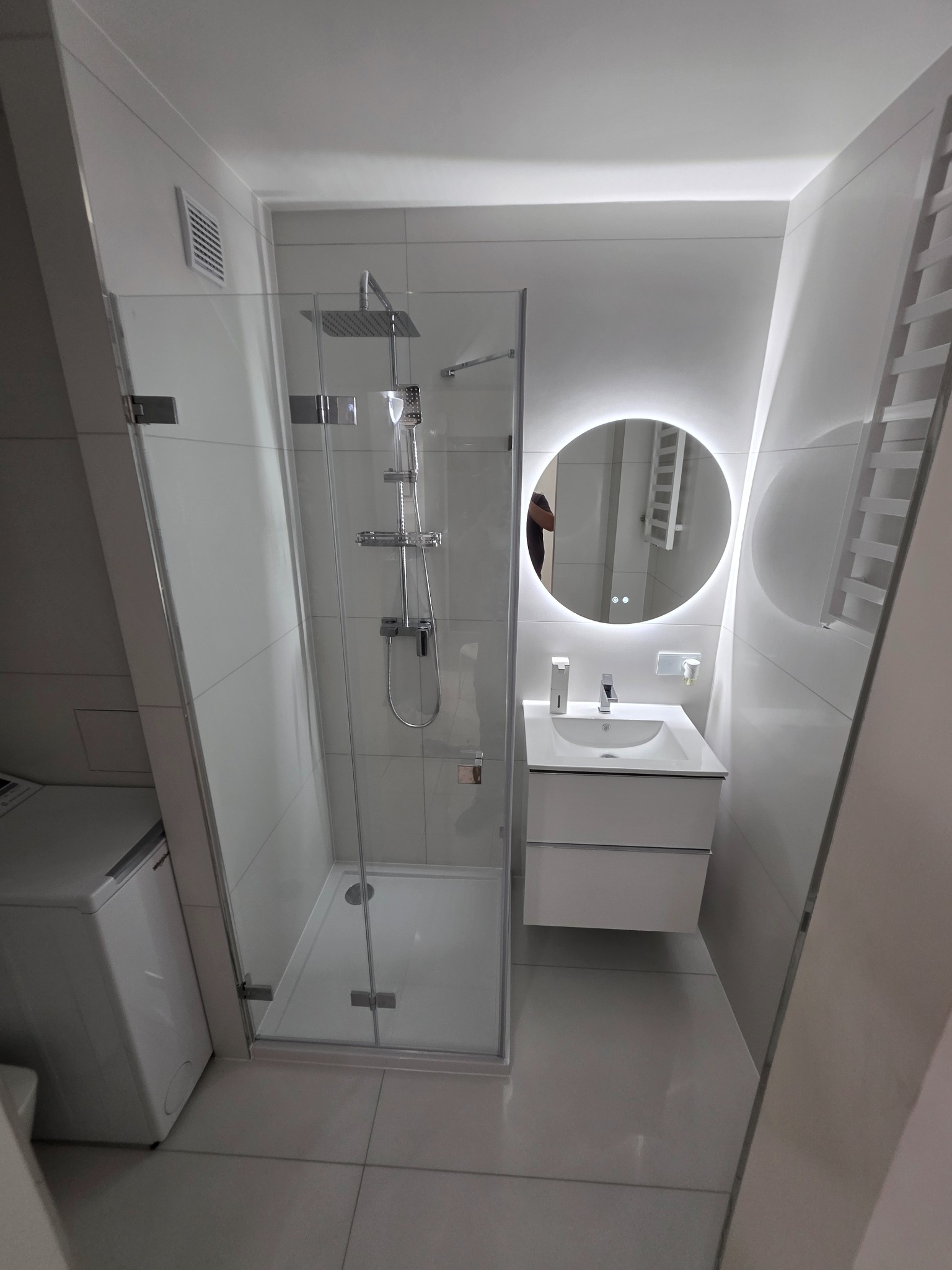 Nowoczesna, biała łazienka z kabiną prysznicową typu walk-in, okrągłym lustrem z podświetleniem LED i umywalką z szafką. Widoczna pralka i grzejnik drabinkowy.