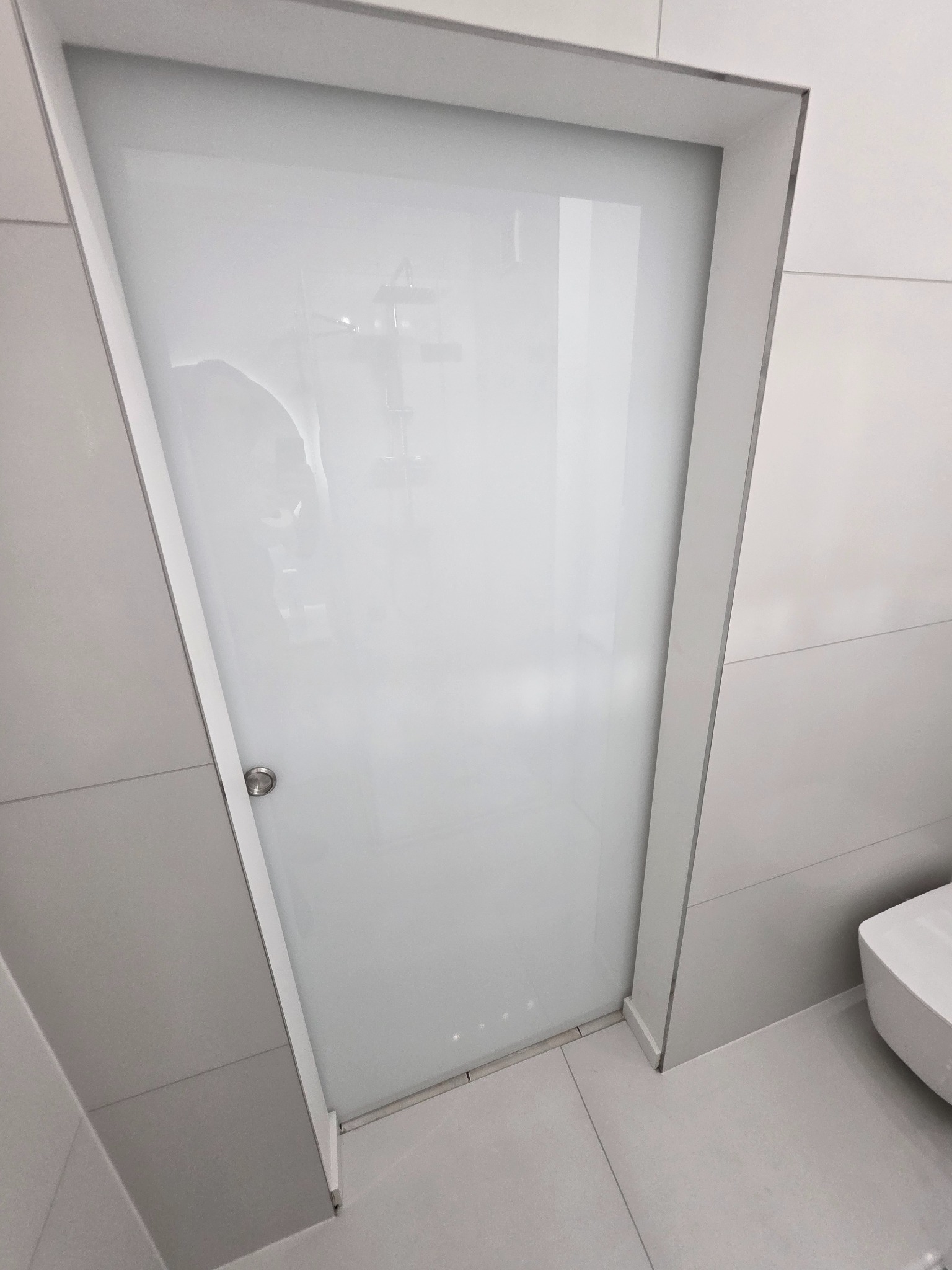 Nowoczesna kabina prysznicowa z matowego szkła, minimalistyczny design, szare płytki, widoczny fragment toalety, jasne wnętrze łazienki po wykończeniu.