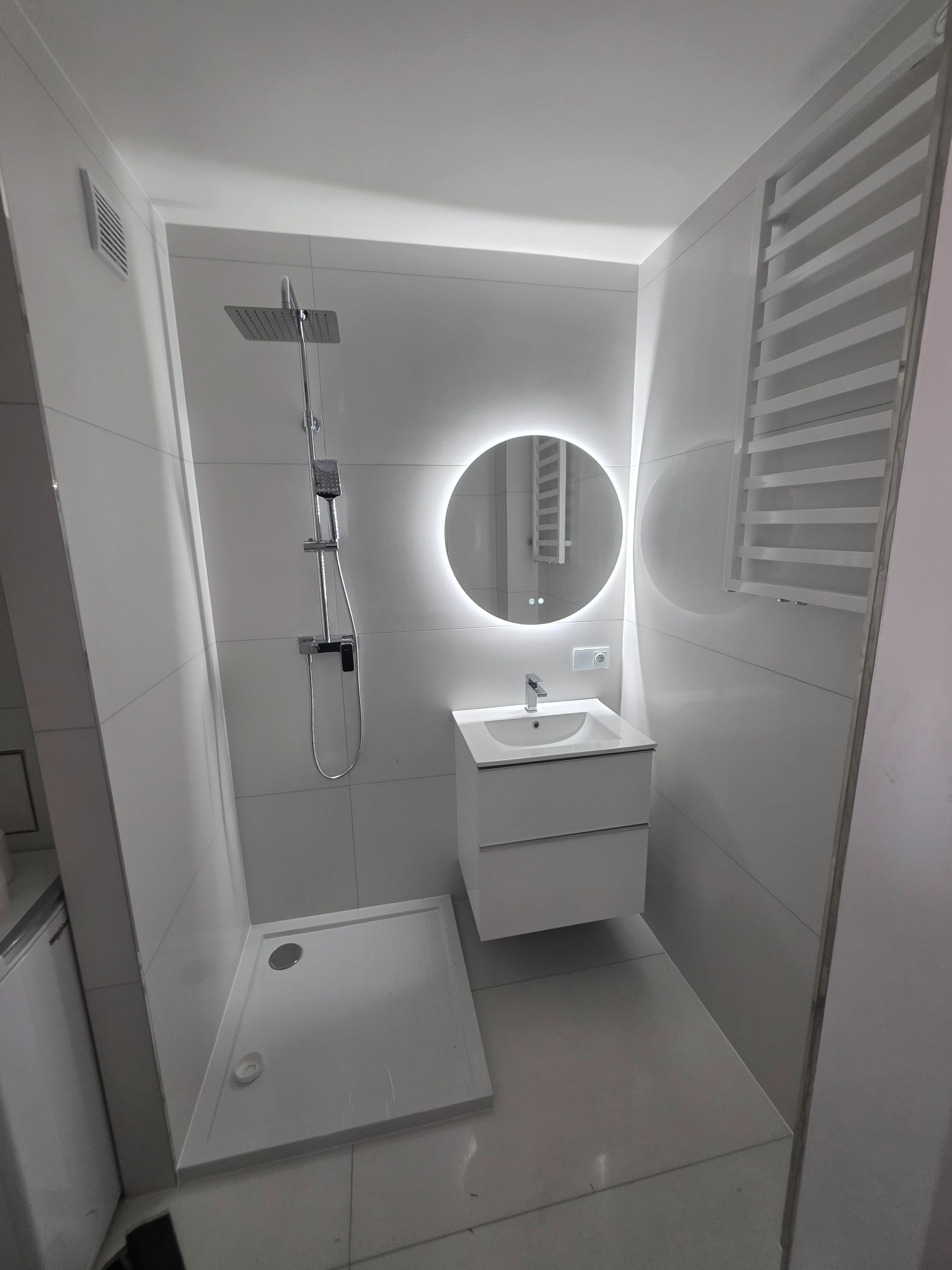 Nowoczesna, biała łazienka z kabiną prysznicową typu walk-in, okrągłym lustrem z podświetleniem LED i grzejnikiem drabinkowym. Minimalistyczny design, jasne płytki.