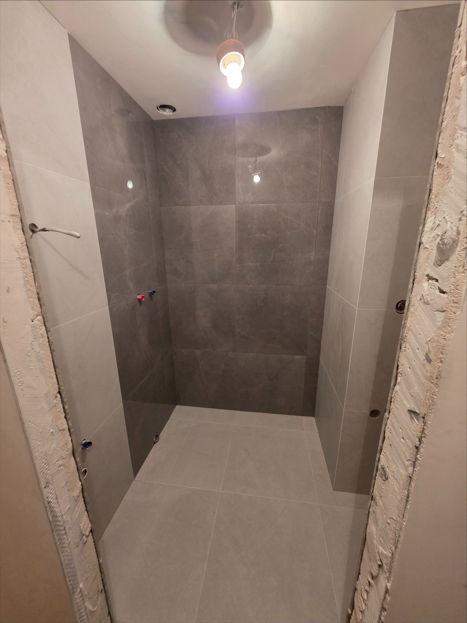 Wykończona kabina prysznicowa z szarymi płytkami imitującymi marmur, widoczne przyłącza wody i surowa ościeżnica. Minimalistyczny design.