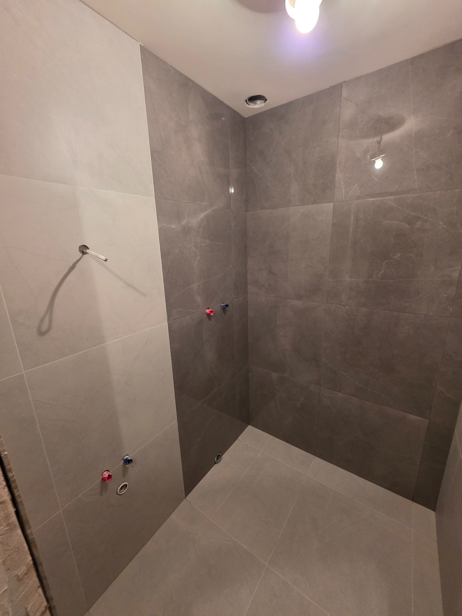 Wykończona łazienka z szarymi płytkami imitującymi marmur na ścianach i podłodze. Widoczne przyłącza wodne i punkt oświetleniowy w suficie.