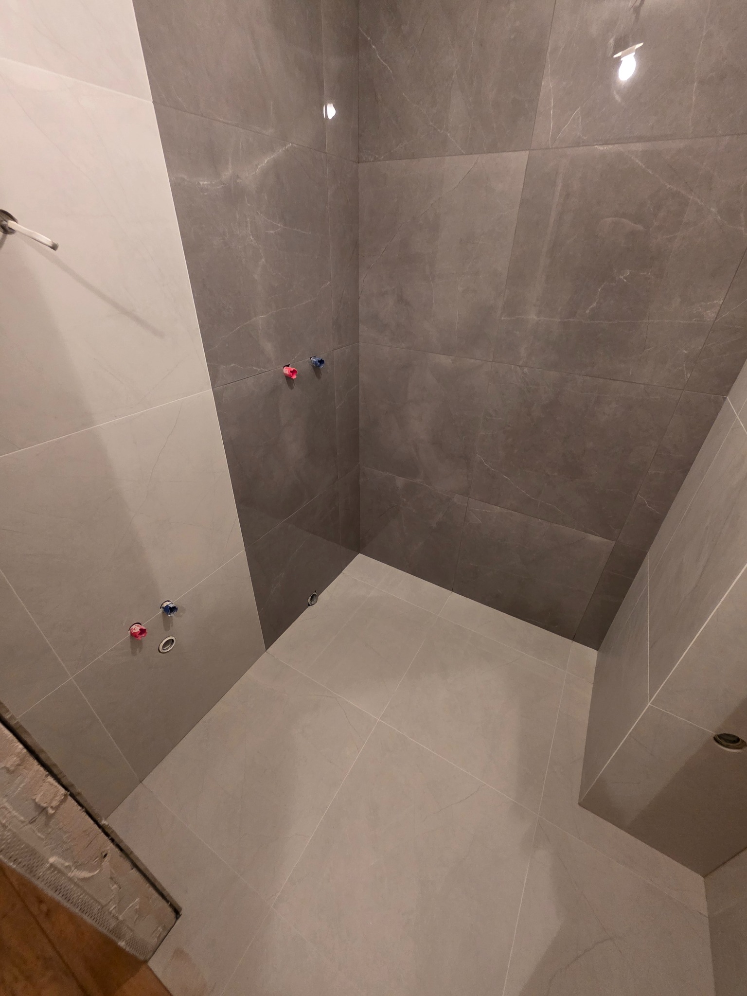 Wykończona łazienka z szarymi płytkami imitującymi marmur na ścianach i podłodze. Widoczne przyłącza wodne i oświetlenie sufitowe. Minimalistyczny design.