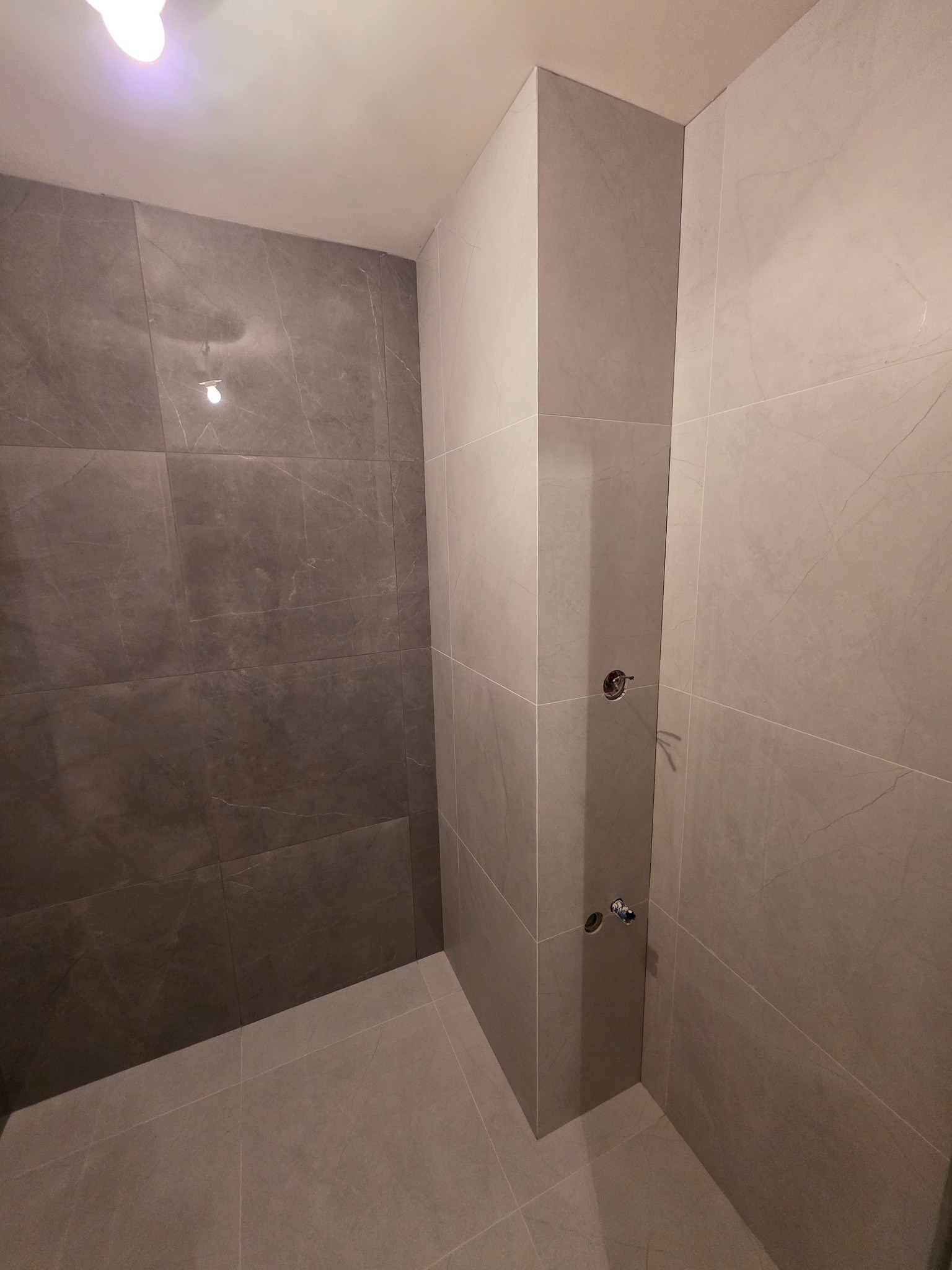 Wykończona łazienka z szarymi płytkami imitującymi marmur. Widoczne przyłącza do baterii. Minimalistyczny design, nowoczesne wykończenie.