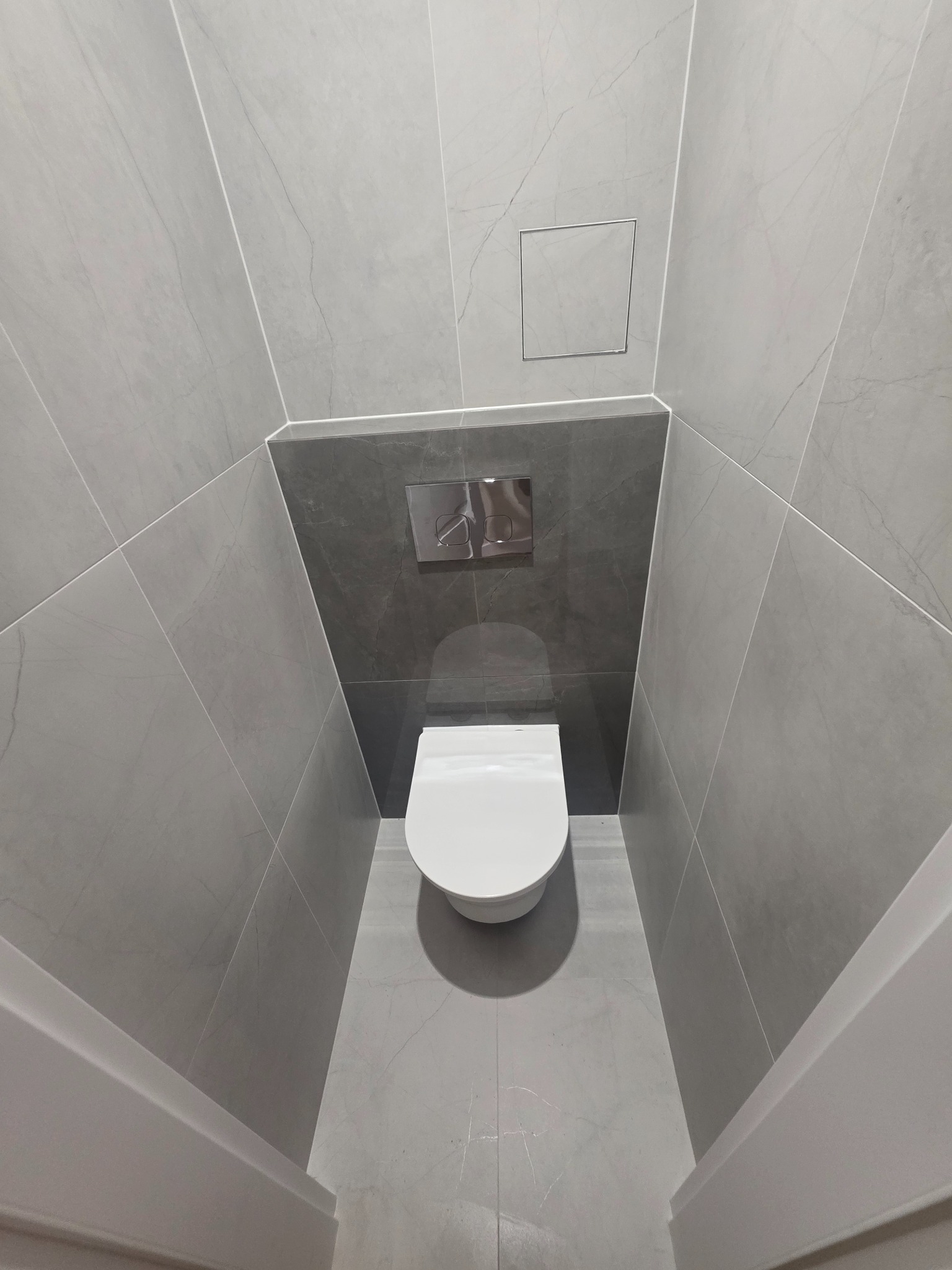Nowoczesna łazienka w szarościach z podwieszaną toaletą i geometrycznym układem płytek. Widoczny chromowany przycisk spłukiwania i ukryty właz rewizyjny.