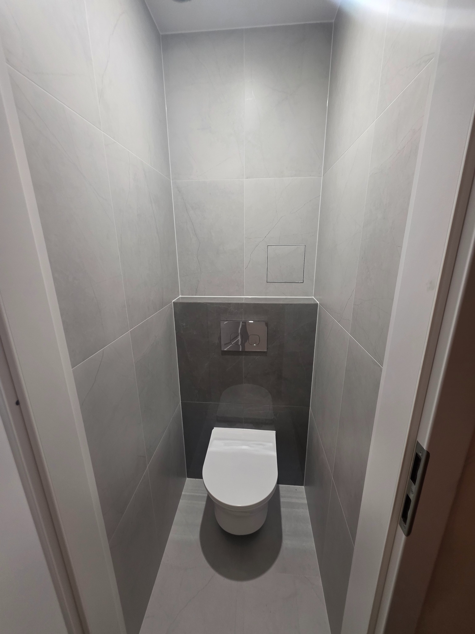 Wąska toaleta w szarościach: płytki imitujące marmur, podwieszana miska WC, chromowany przycisk spłukujący. Minimalistyczny design, nowoczesne wykończenie.