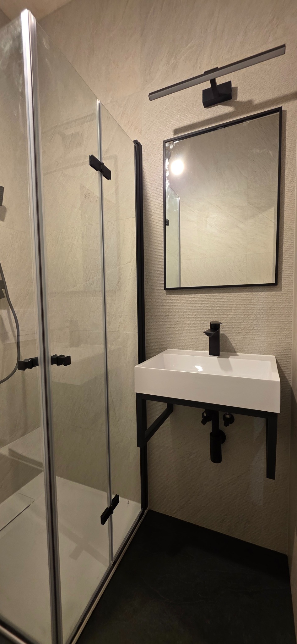 Nowoczesna łazienka z kabiną prysznicową, białą umywalką na czarnym stelażu i lustrem z oświetleniem. Beżowe płytki na ścianach i ciemna podłoga.