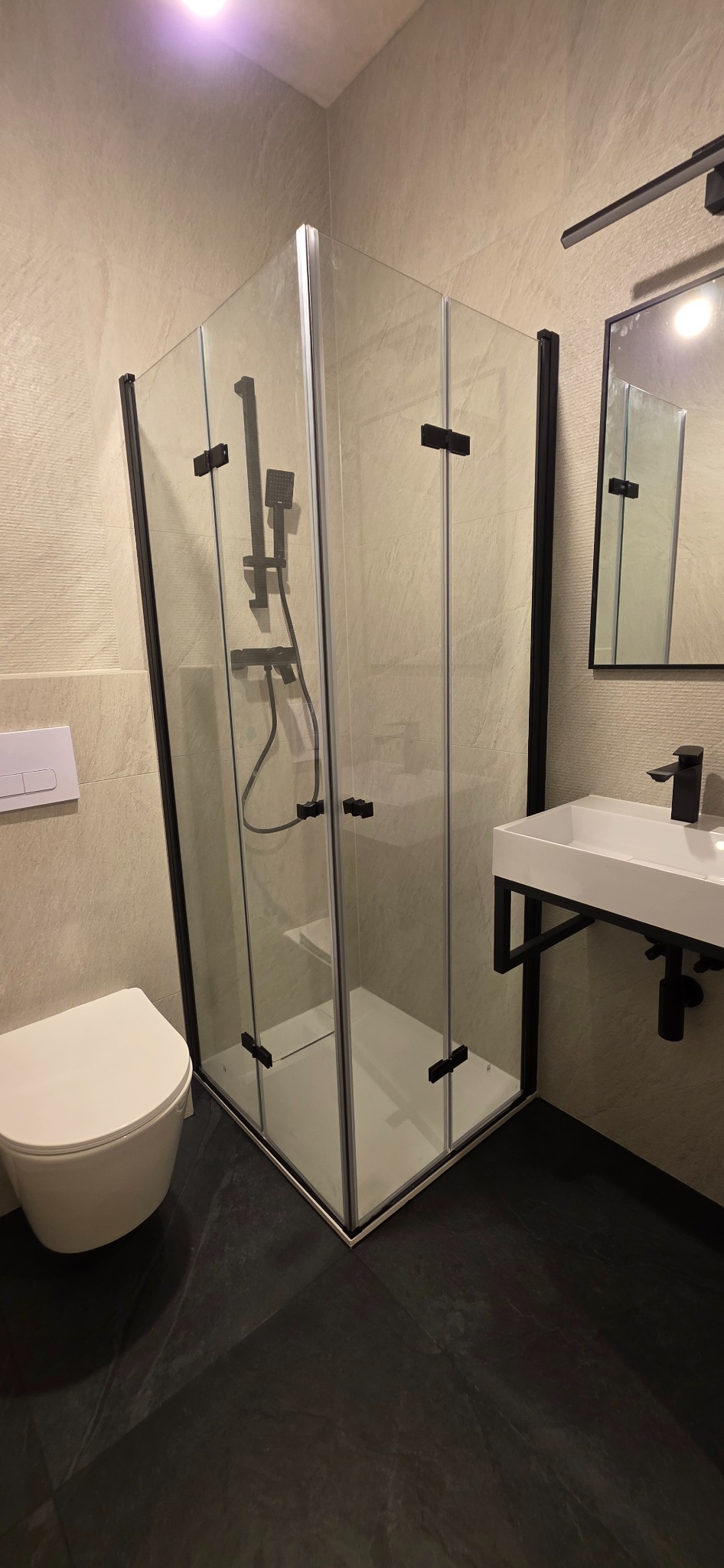 Nowoczesna łazienka z kabiną prysznicową typu walk-in z czarnymi profilami, białą umywalką i toaletą. Ciemna podłoga i jasne płytki na ścianach.