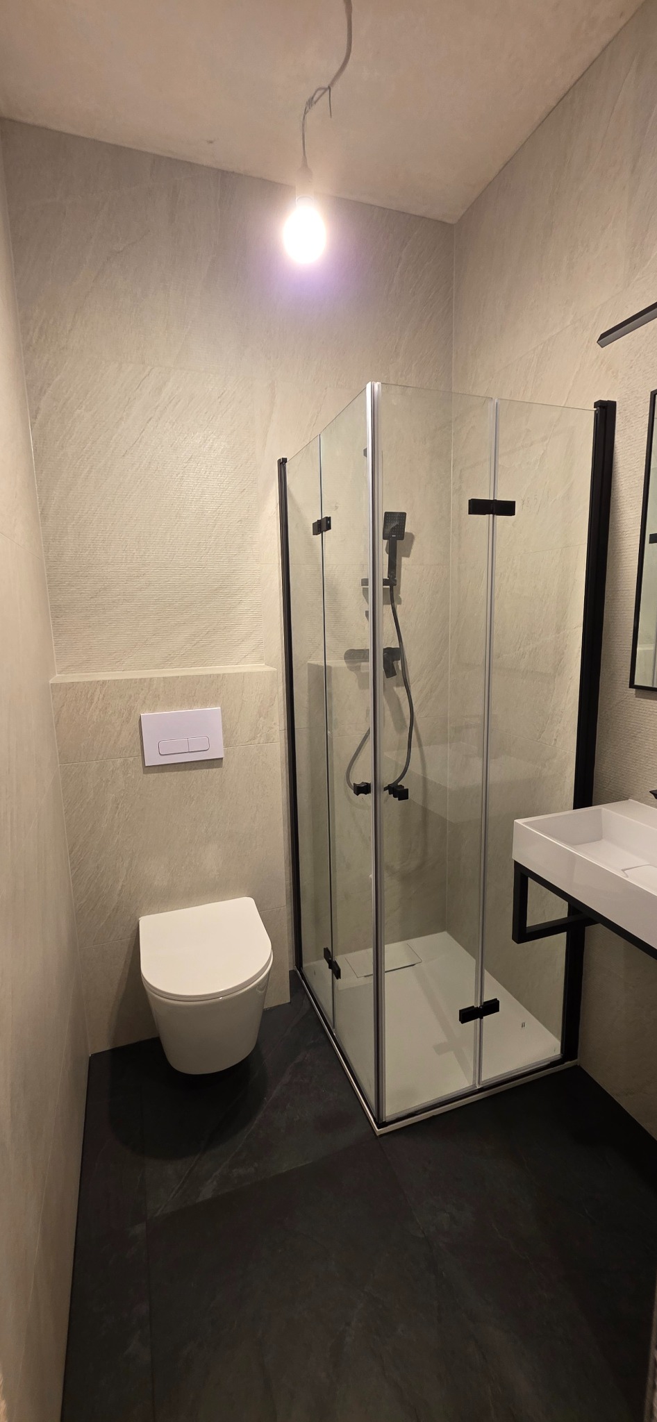Nowoczesna łazienka z kabiną prysznicową typu walk-in, podwieszaną toaletą i umywalką na czarnym stelażu. Ściany wykończone płytkami w odcieniach beżu, podłoga w ciemnym kolorze.