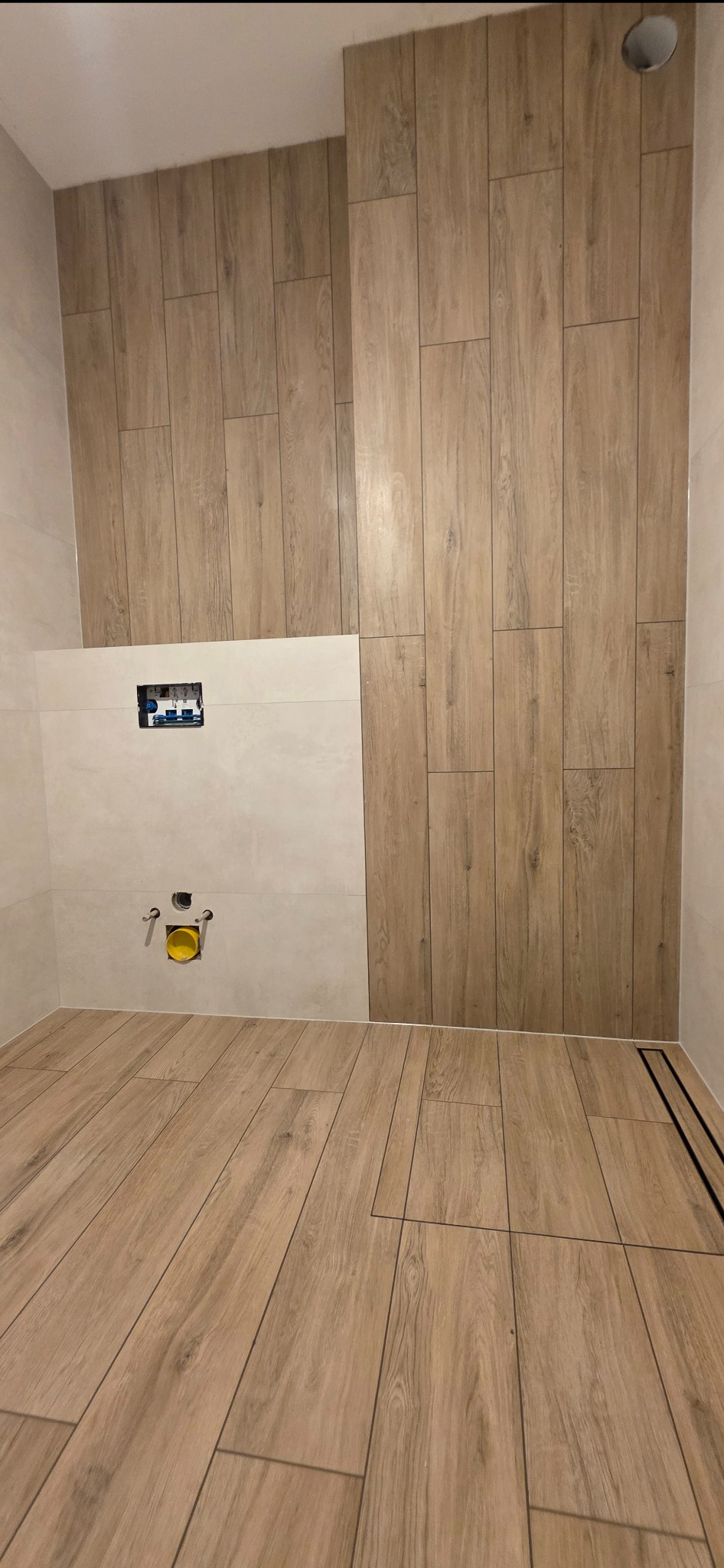 Wykończona łazienka z drewnopodobnymi płytkami na podłodze i ścianie. Widoczny odpływ liniowy i instalacja podtynkowa WC. Jasne, ciepłe oświetlenie.