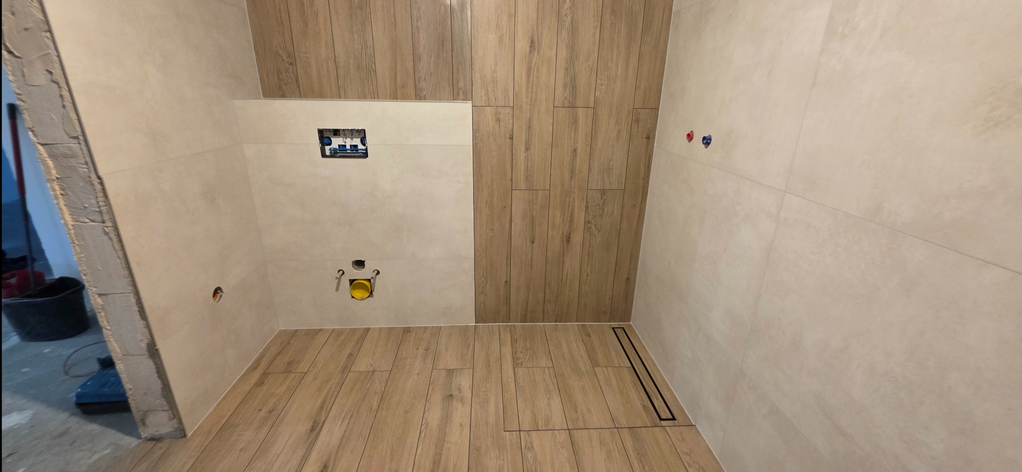 Wykończona łazienka z beżowymi płytkami imitującymi drewno na podłodze i części ściany. Widoczny odpływ liniowy i instalacja podtynkowa WC.