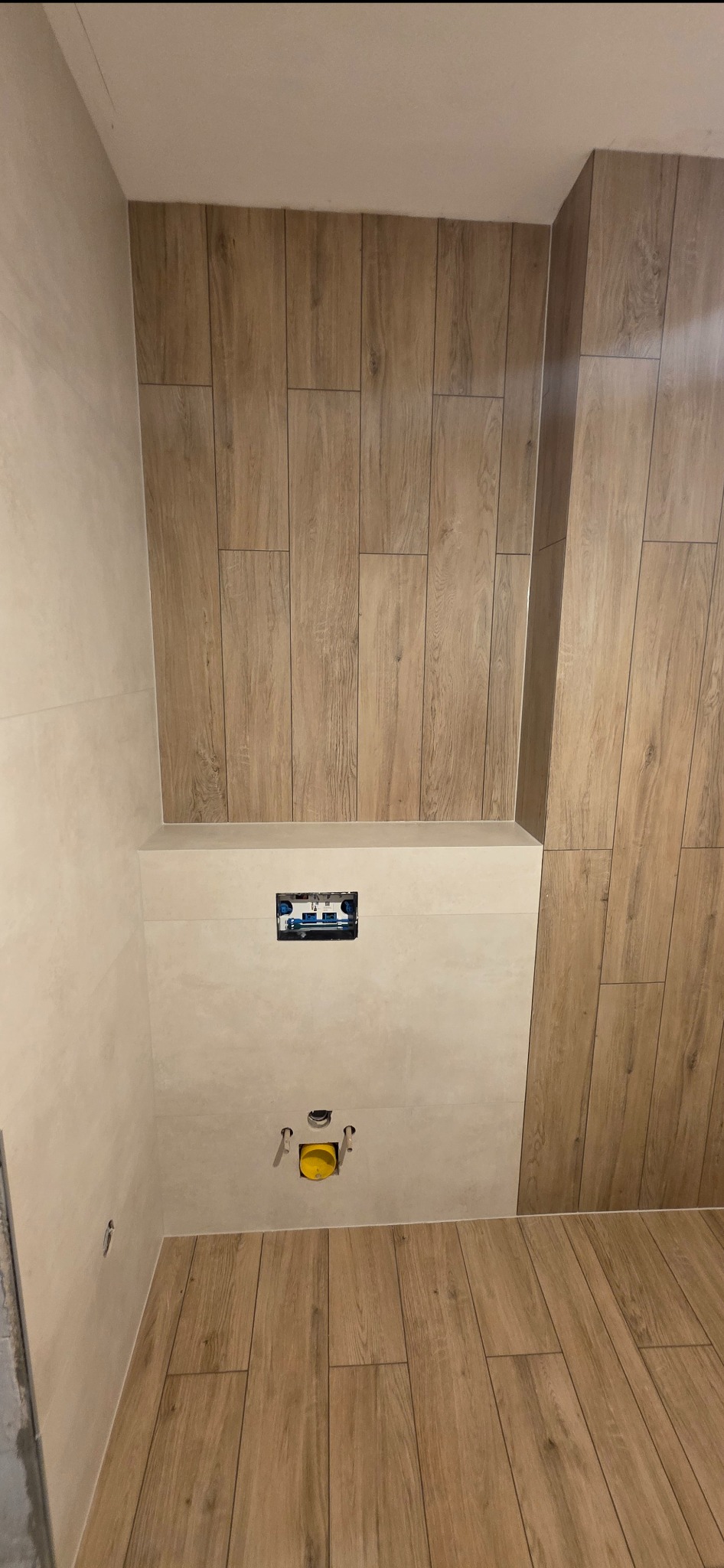 Wykończona łazienka z drewnopodobnymi płytkami na ścianach i podłodze. Widoczne podłączenia do toalety podtynkowej i puszka elektryczna. Jasne, neutralne barwy.