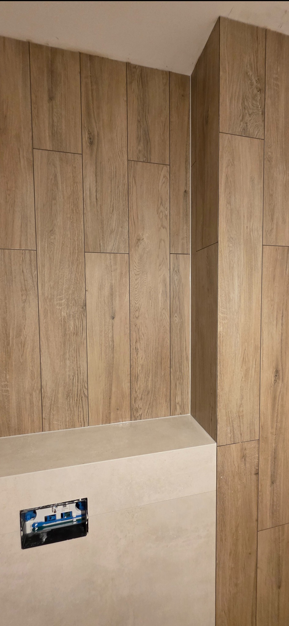 Wykończona ściana w łazience z płytek imitujących drewno, z widocznym fragmentem zabudowy geberitu. Minimalistyczny design, ciepłe odcienie brązu i beżu.