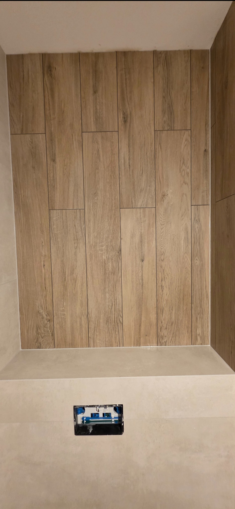 Wykończona ściana w łazience z płytek imitujących drewno, z widocznym otworem na armaturę. Jasne, neutralne barwy, minimalistyczny design.