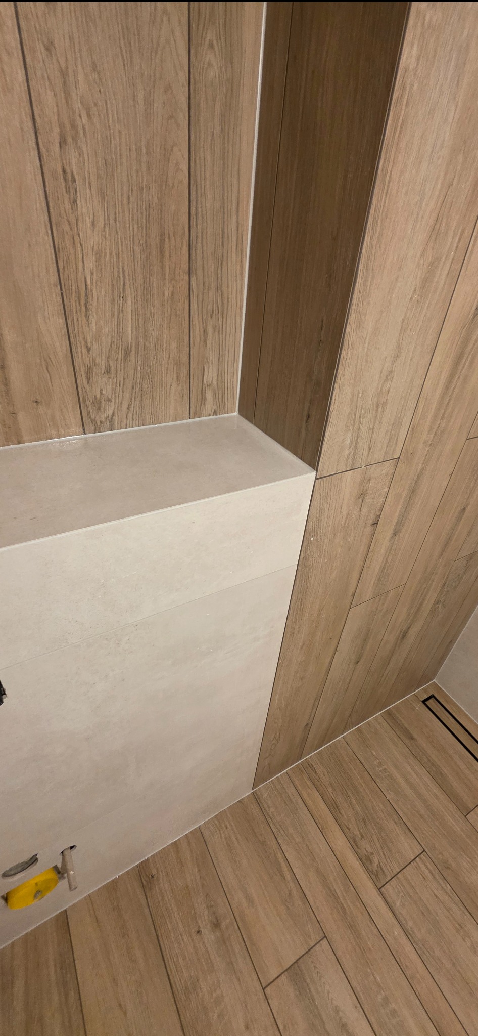 Nowoczesna łazienka z drewnopodobnymi płytkami na ścianach i podłodze, jasny blat. Widoczny odpływ liniowy i narzędzie glazurnicze. Minimalistyczny design.