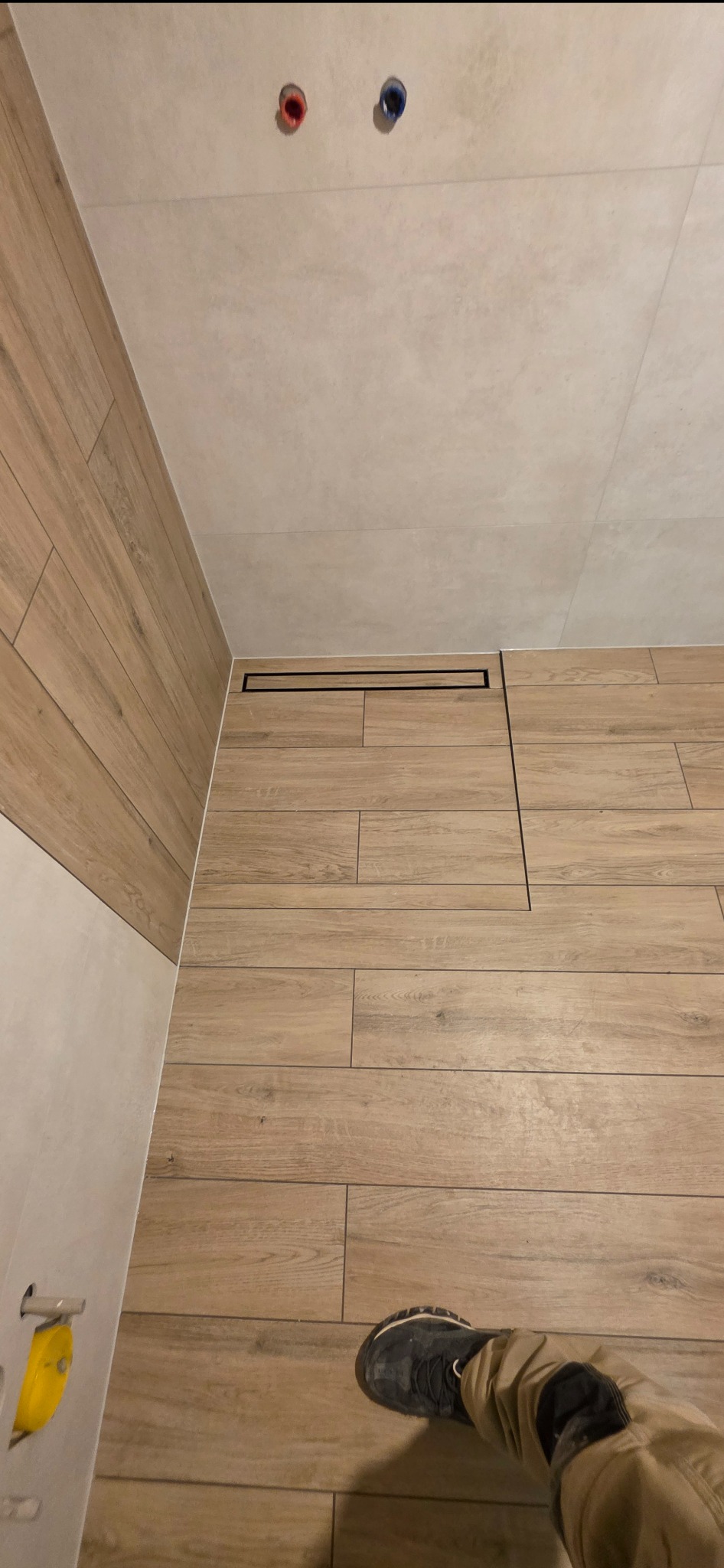 Wykończona łazienka z drewnopodobnymi płytkami, czarnym odpływem liniowym i widocznymi przyłączami wodnymi. Ujęcie z perspektywy osoby stojącej w rogu pomieszczenia.