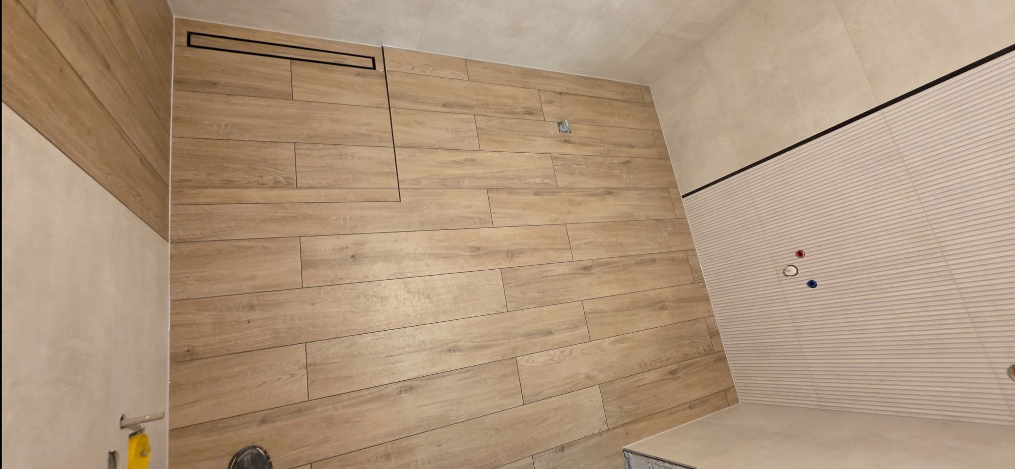 Wykończona ściana łazienki z drewnopodobnych płytek, z widocznym odpływem liniowym i fragmentem ściany z białych, ryflowanych paneli. Widoczne przyłącza wodne.