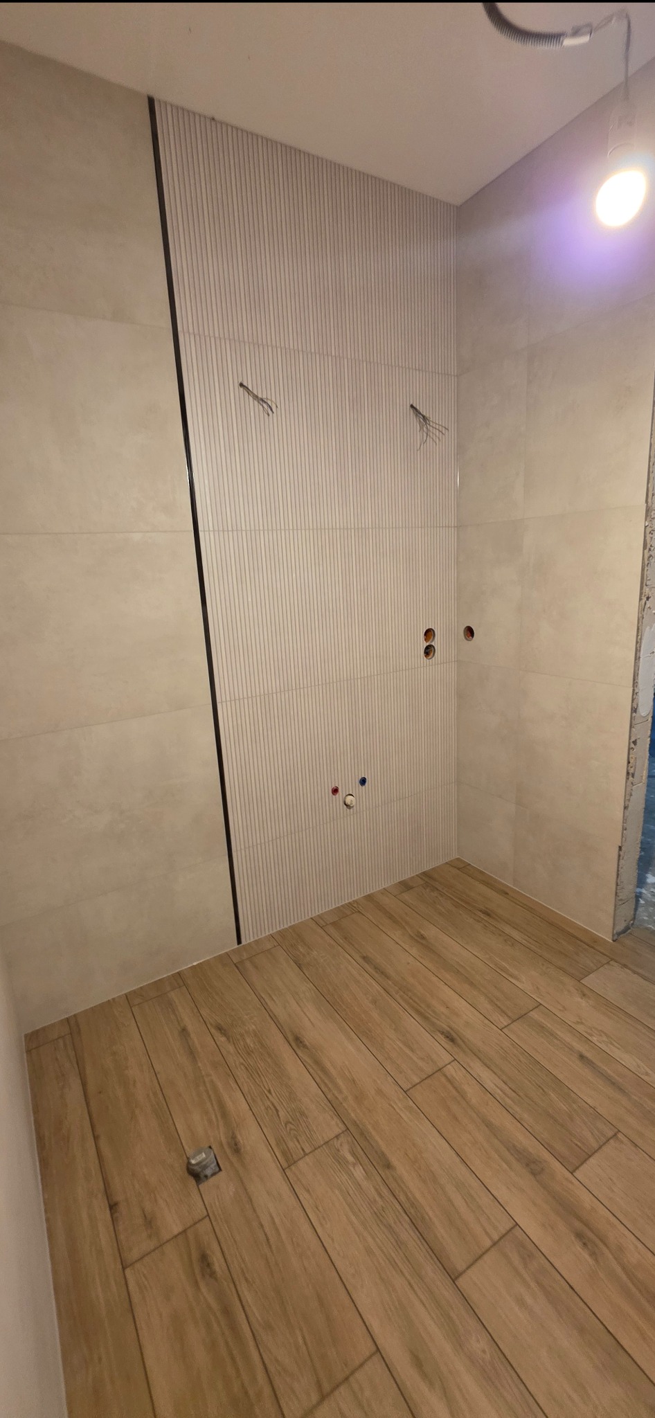 Wykończona łazienka z beżowymi płytkami imitującymi drewno na podłodze i ścianach. Widoczne wystające kable i rury, sugerujące etap prac wykończeniowych.