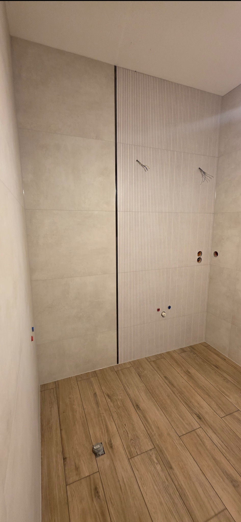 Wykończona łazienka z drewnopodobną podłogą i płytkami w odcieniach beżu. Widoczne przygotowane punkty instalacji wodnej i elektrycznej. Minimalistyczny design.