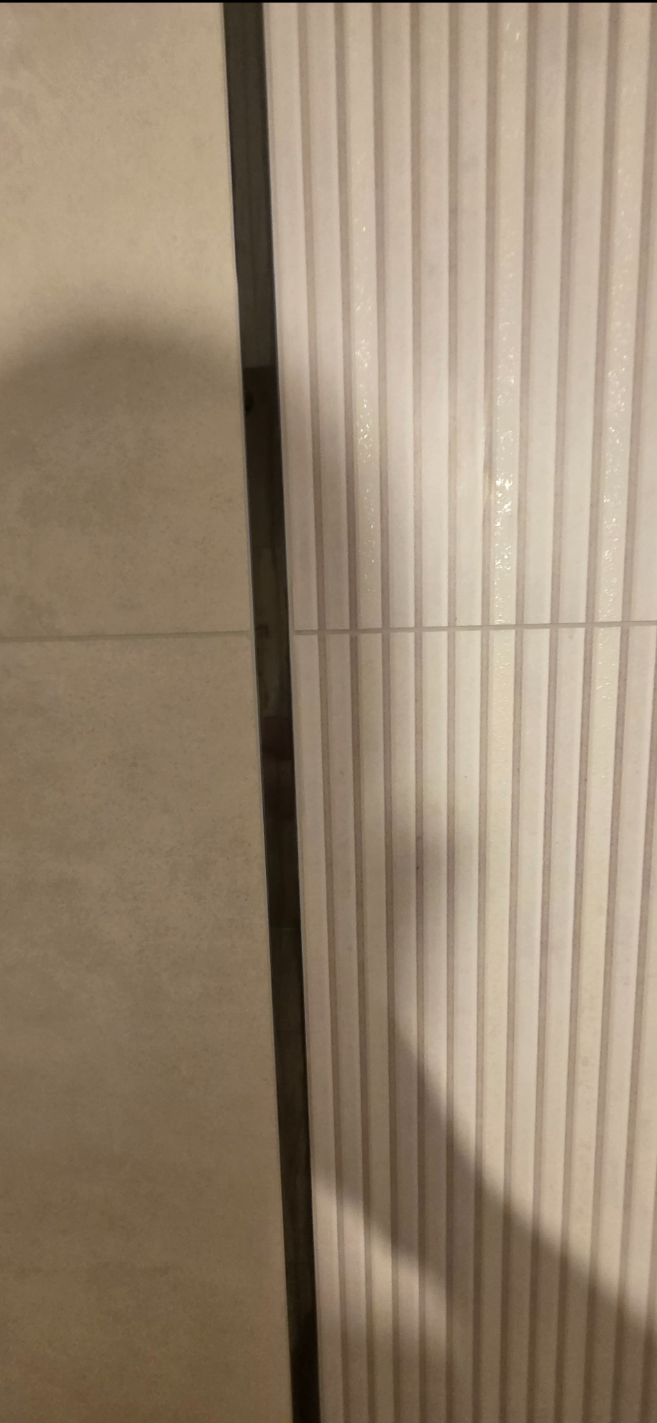 Beżowe płytki ścienne z delikatnym wzorem pionowych pasków, oddzielone ciemną listwą. Widoczny fragment ściany po remoncie, detale wykończenia.