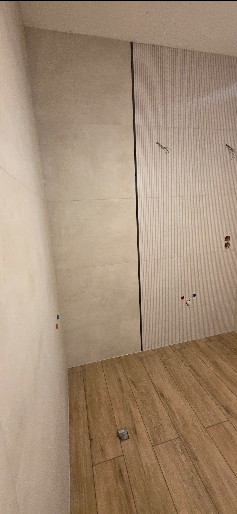 Wykończona łazienka z beżowymi płytkami na ścianach, w tym jedna ściana z pionowymi paskami. Na podłodze drewnopodobne płytki. Widoczne wystające kable i rury.