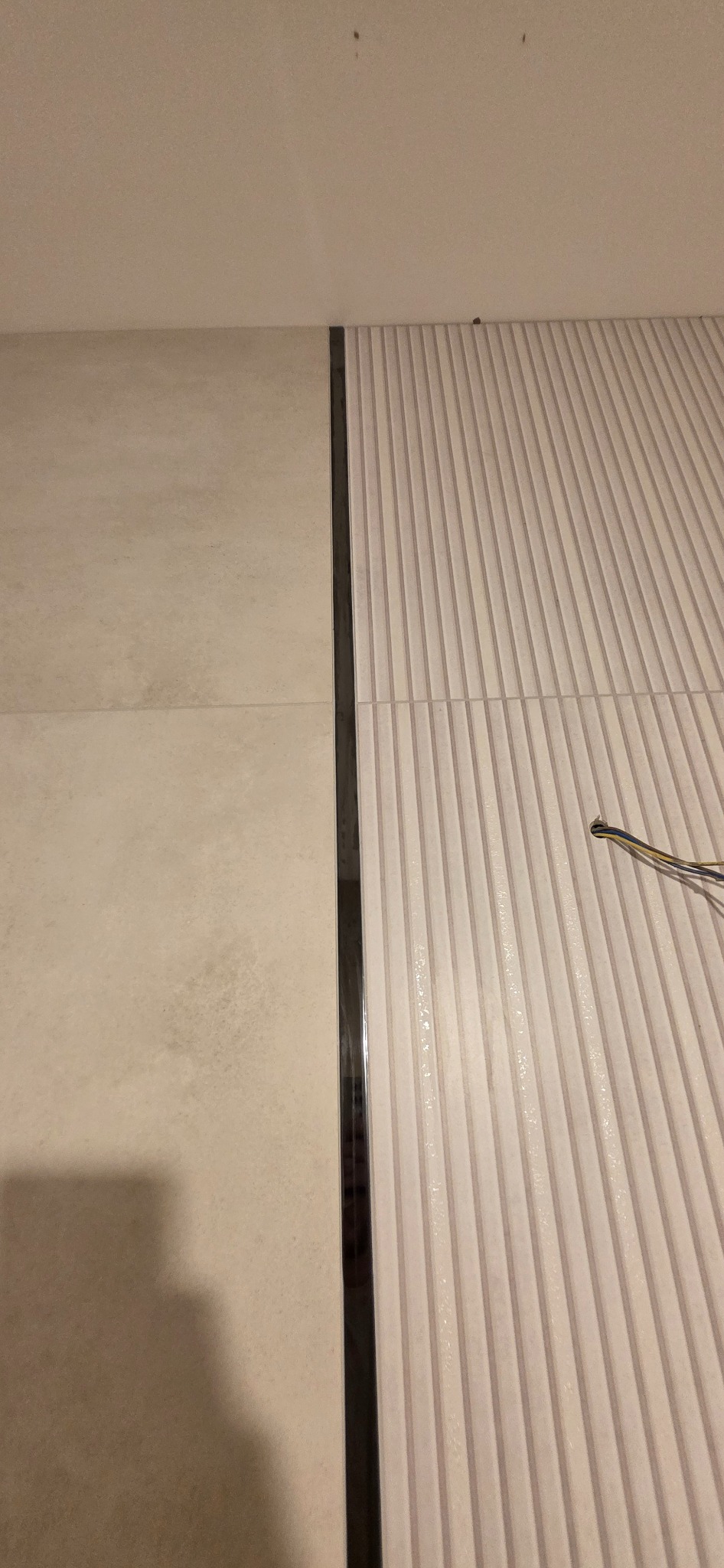 Ułożone płytki podłogowe w łazience, z widocznym odpływem liniowym i fragmentem instalacji elektrycznej. Płytki w dwóch odcieniach beżu, z fakturą.