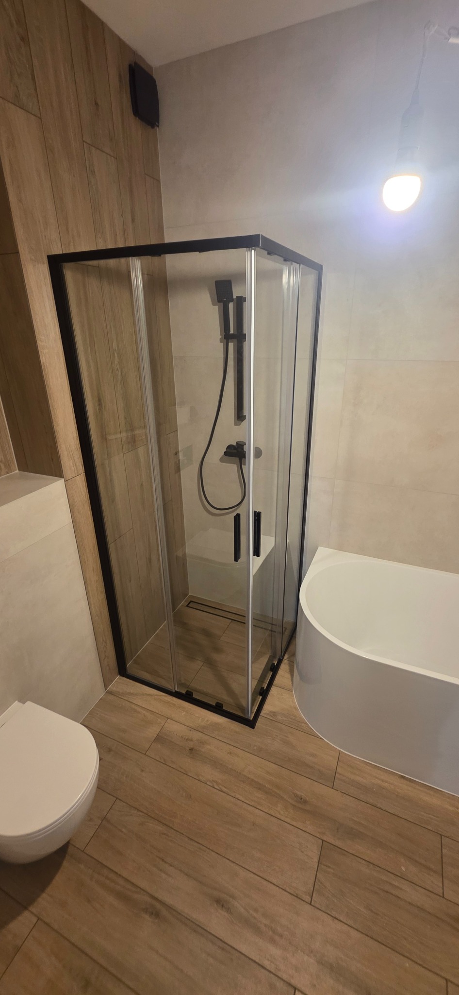 Nowoczesna łazienka z kabiną prysznicową typu walk-in z czarnymi profilami, białą wanną i toaletą. Płytki imitujące drewno na podłodze i ścianach.