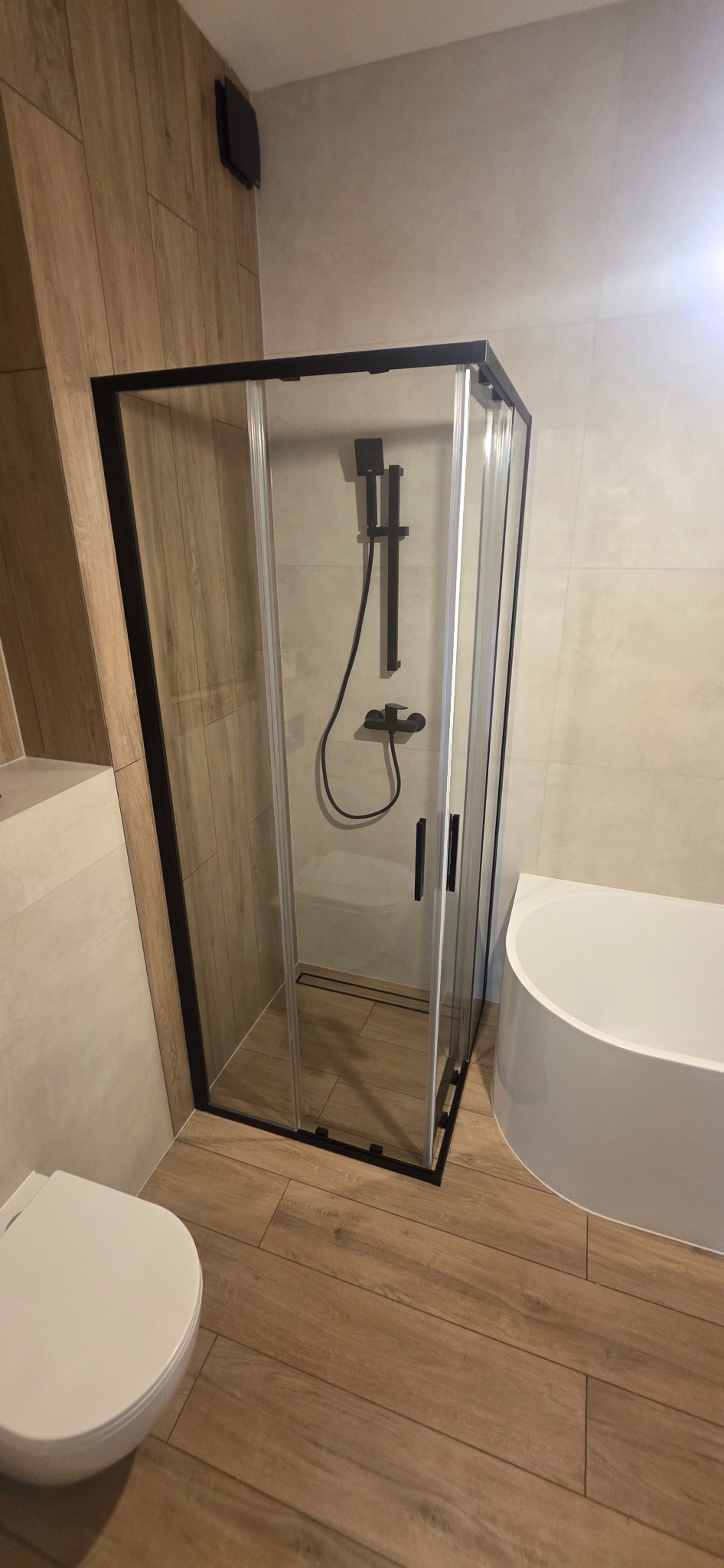 Nowoczesna kabina prysznicowa z czarnymi profilami i deszczownicą w wykończonej łazience z drewnopodobnymi płytkami. Widoczna toaleta i fragment wanny.
