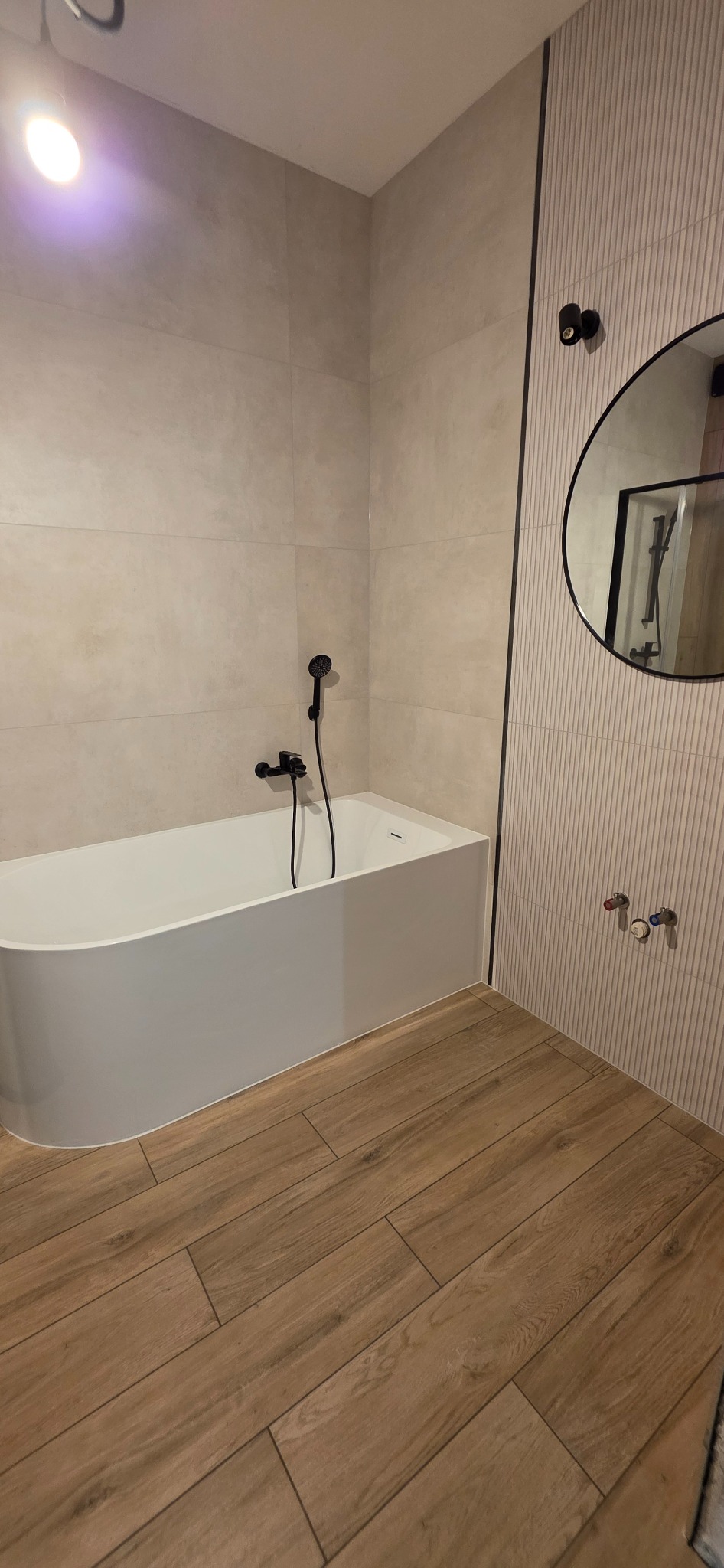 Nowoczesna łazienka z wolnostojącą wanną, czarną armaturą i drewnopodobną podłogą. Na ścianie okrągłe lustro z czarną ramą i dekoracyjne pionowe panele.