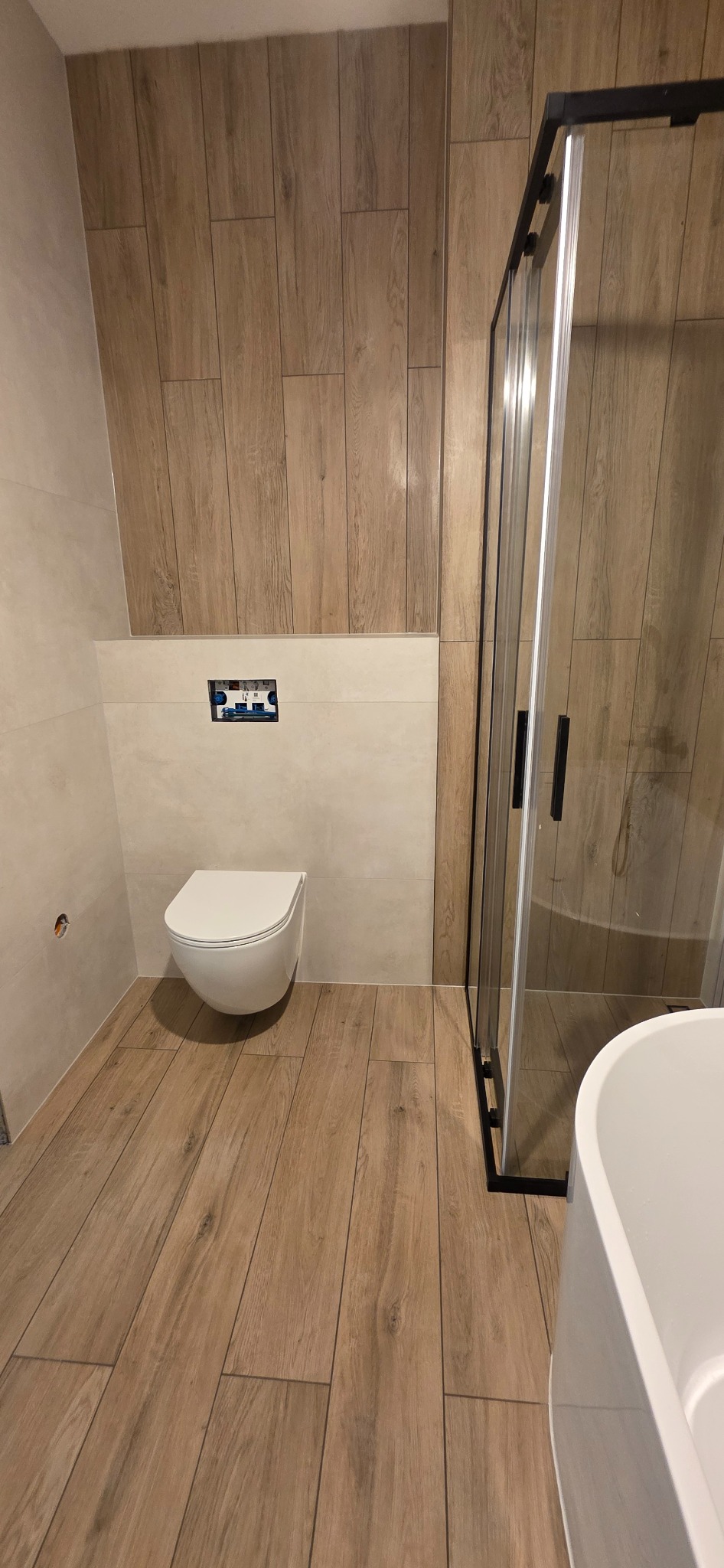 Nowoczesna łazienka z drewnopodobnymi płytkami, podwieszaną toaletą i kabiną prysznicową z czarnymi profilami. Minimalistyczny design, jasne kolory, ciepłe oświetlenie.